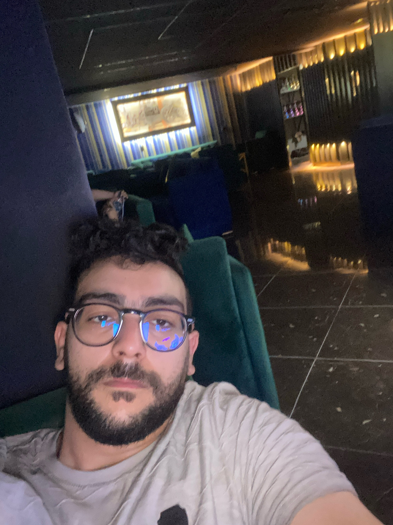 A medium shot selfie shows a man with dark, curly hair and a beard, wearing glasses, and a light-colored t-shirt. He is seated in what appears to be a lounge or dimly lit seating area. The background reveals a modern interior with dark furniture, striped curtains in blue and yellow, and illuminated wall panels. A framed picture hangs on the wall behind the curtains. In the distance, a bar or shelf area with bottles is visible, also lit from above. The floor is dark and reflective, showing the overhead lighting. The overall atmosphere suggests a relaxed evening or night setting in a city establishment. Given the prompt's request to place this in Meknes, Morocco, it is likely a sophisticated lounge or bar within the city.