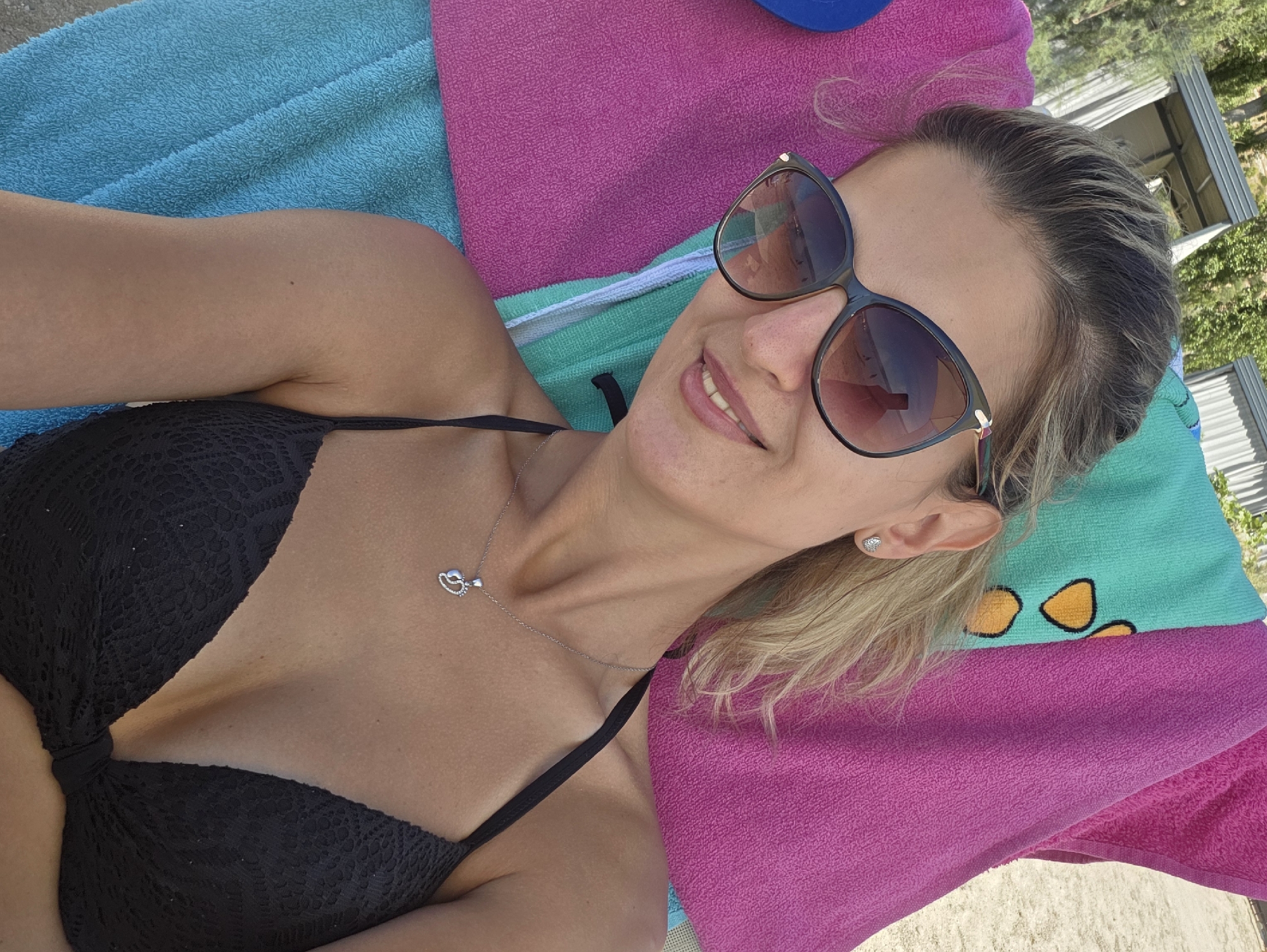 A woman with blonde hair, wearing sunglasses and a black bikini top, lies on a beach chair covered with brightly colored towels. She is smiling and looking at the camera. The background shows a sandy beach, the sea, and some trees. The scene appears to be in Omiš, Croatia, possibly near the Cetina river mouth, given the typical beach scenery of the area.