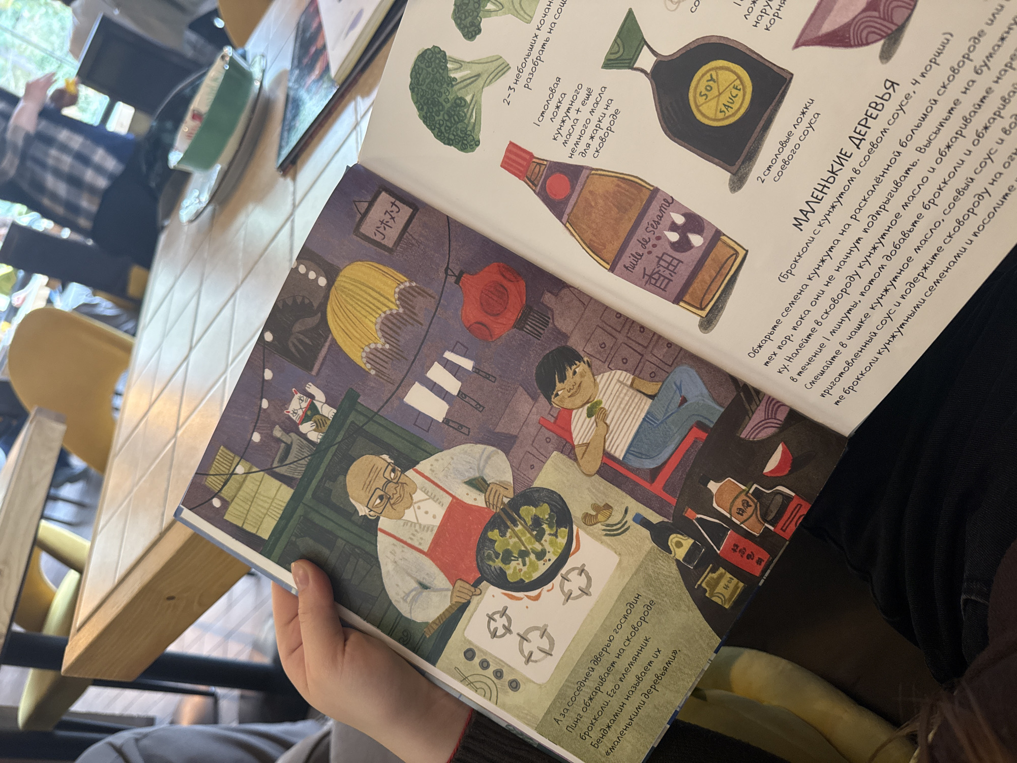 The image captures an indoor scene within a cafe or restaurant in Sochi, Russia. In the immediate foreground, a person's hands, with manicured nails, hold open an illustrated book. The book displays a colorful two-page spread. The left page features an illustration of an elderly Asian man, wearing glasses and a red apron, actively cooking broccoli in a black wok on a stovetop. To his right, a younger Asian boy sits on a red chair, holding and seemingly eating a piece of broccoli. Below them, various bottles, including a dark one labeled "Huile de Sésame" and another resembling soy sauce, are depicted on a surface next to a small red bowl. The background of this illustration includes hanging red lanterns and a white Maneki-neko cat figurine. Russian text is visible on the lower left portion of this page, describing the illustrated scene.
The right page of the book features illustrations of two broccoli florets, a tall bottle labeled "Huile de Sésame" (sesame oil) with Chinese characters, and a black bottle marked "SOY SAUCE" with a yellow label. Below these illustrations, Russian text outlines a recipe titled "МАЛЕНЬКИЕ ДЕРЕВЬЯ" (Little Trees), detailing ingredients and cooking instructions for broccoli with sesame and soy sauce.
The background of the photograph is out of focus, revealing light-colored wooden tables with square tiled tops and yellow-backed chairs. Several blurry figures of other individuals are visible in the mid-ground and background, including one person wearing a plaid shirt. A green and white bowl rests on one of the tables in the distance. The overall lighting suggests natural light entering the establishment.