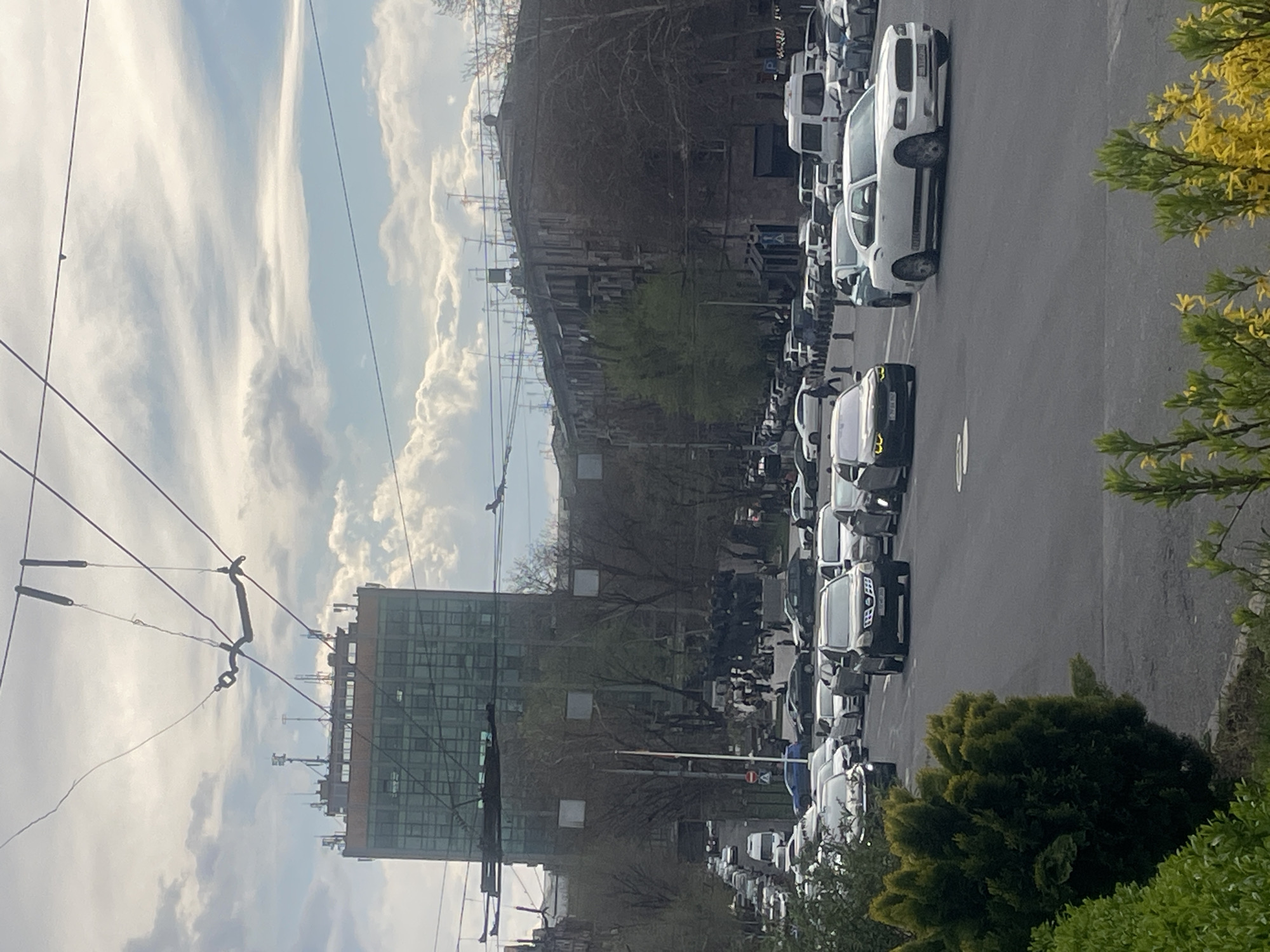 The image captures a street in Yerevan, Armenia, lined with a large number of white delivery vans parked along the right side of the road. A modern glass building is visible on the left, along with a complex of power lines and telecommunication antennas. In the background, a large, old industrial building is situated on a slight hill. The sky is partly cloudy with patches of blue. The scene appears to be taken from an elevated position, with trees and greenery partially framing the foreground on the right.
