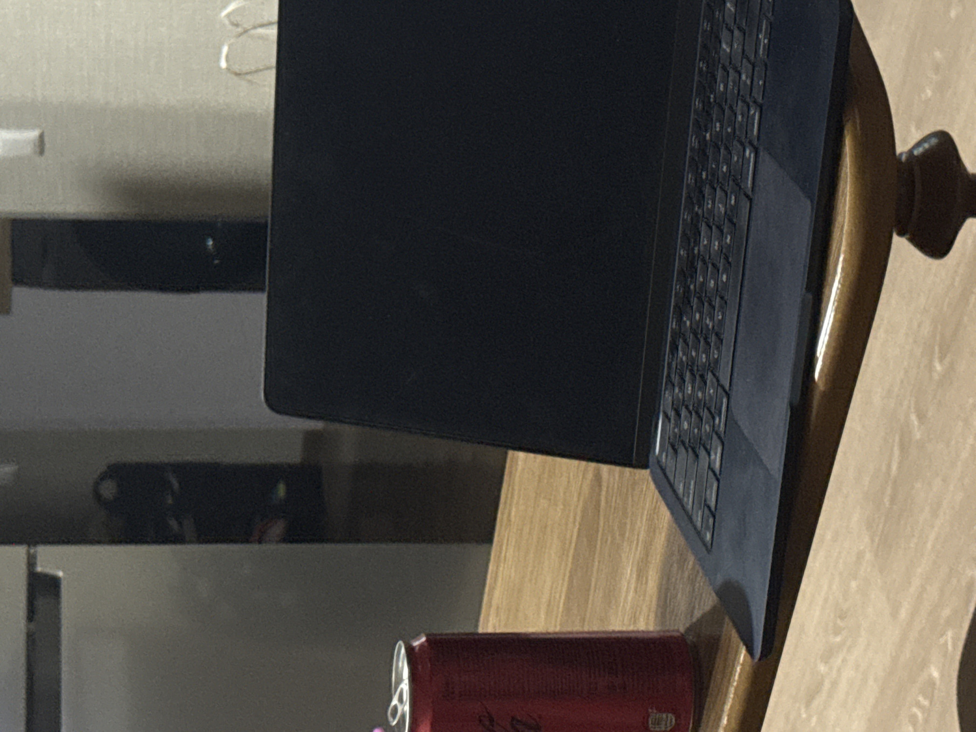 The image shows a close-up view of a dark-colored laptop computer placed on a wooden table. A red can of soda is positioned to the left of the laptop, with its pull-tab visible at the top. The background is blurred, but appears to be an indoor setting, possibly a room with a refrigerator or cabinet to the left and a wall with a light fixture to the right. The lighting suggests it is daytime, but the room may not be well-lit. There are no people visible in the image.