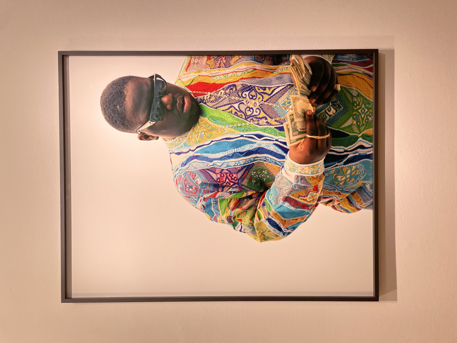 A framed portrait of Notorious B.I.G. wearing sunglasses and holding a wad of cash is on display. The artwork features a white background with a focused image of the rapper in a colorful sweater, gold chain, and multiple rubber bands on his fingers, emphasizing his iconic style. The black frame contrasts with the art, and the portrait hangs on a wall with a neutral, light orange hue.