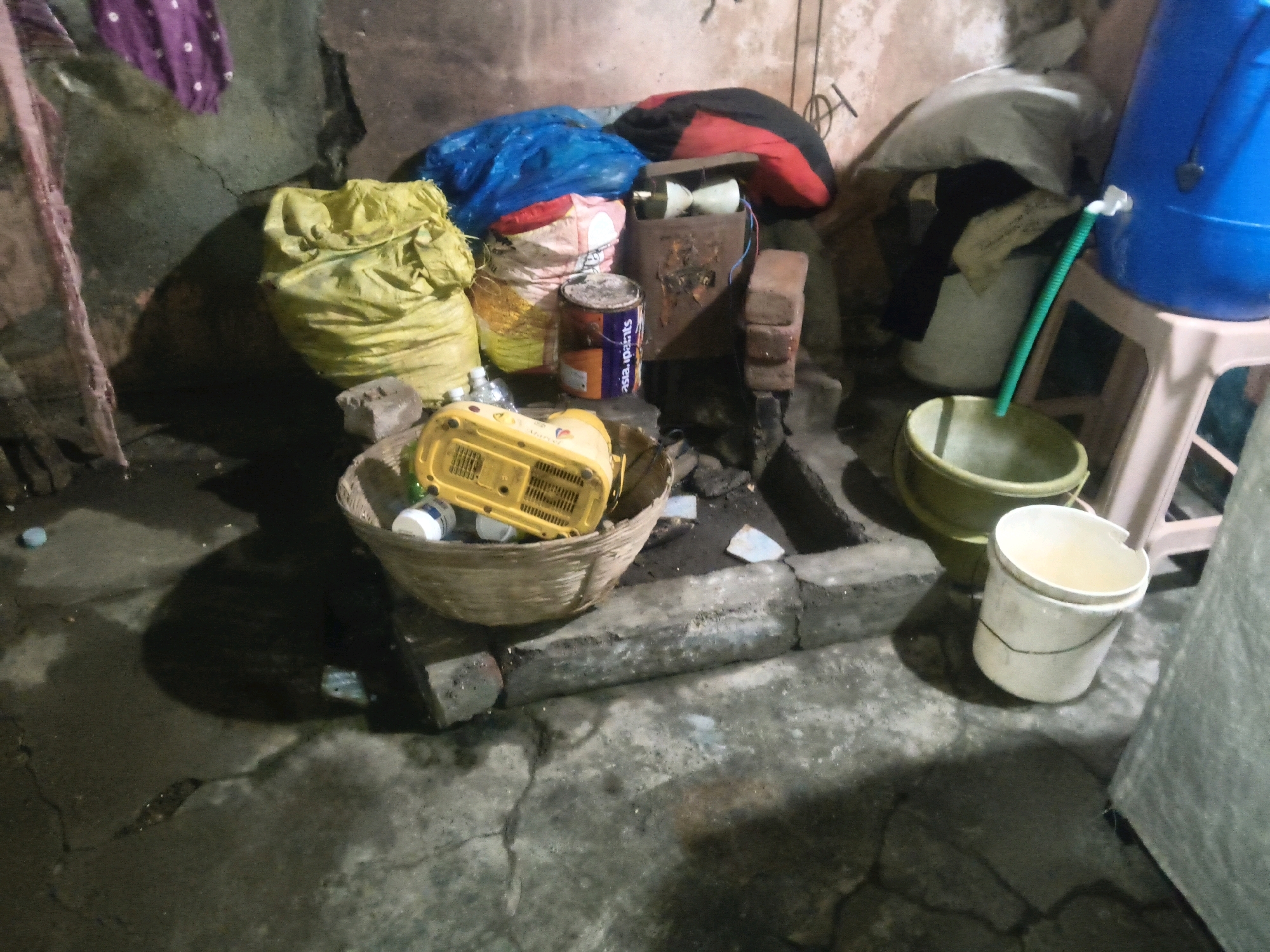 This image depicts a cluttered outdoor or semi-outdoor space, possibly a storage area or a less-maintained yard, located in Hathoda, India. In the foreground, a cracked concrete floor is visible, with shadows cast by various objects. To the left, a large yellow bag is prominent, and next to it, a woven basket contains a yellow electrical appliance, possibly a fan or blower, and some bottles. Behind the basket, several other bags, including one blue and one red and black, are piled together. A tin can with the brand name "asia paints" is also visible among these bags. To the right of the scene, a large blue plastic container is elevated on a stool, with a green tube extending from it into a light green bucket. Another white bucket is placed in front of the green bucket. The background shows a rough, textured wall and various discarded items, suggesting a utilitarian or storage-focused environment. The overall impression is one of disarray and practical necessity rather than aesthetic appeal.