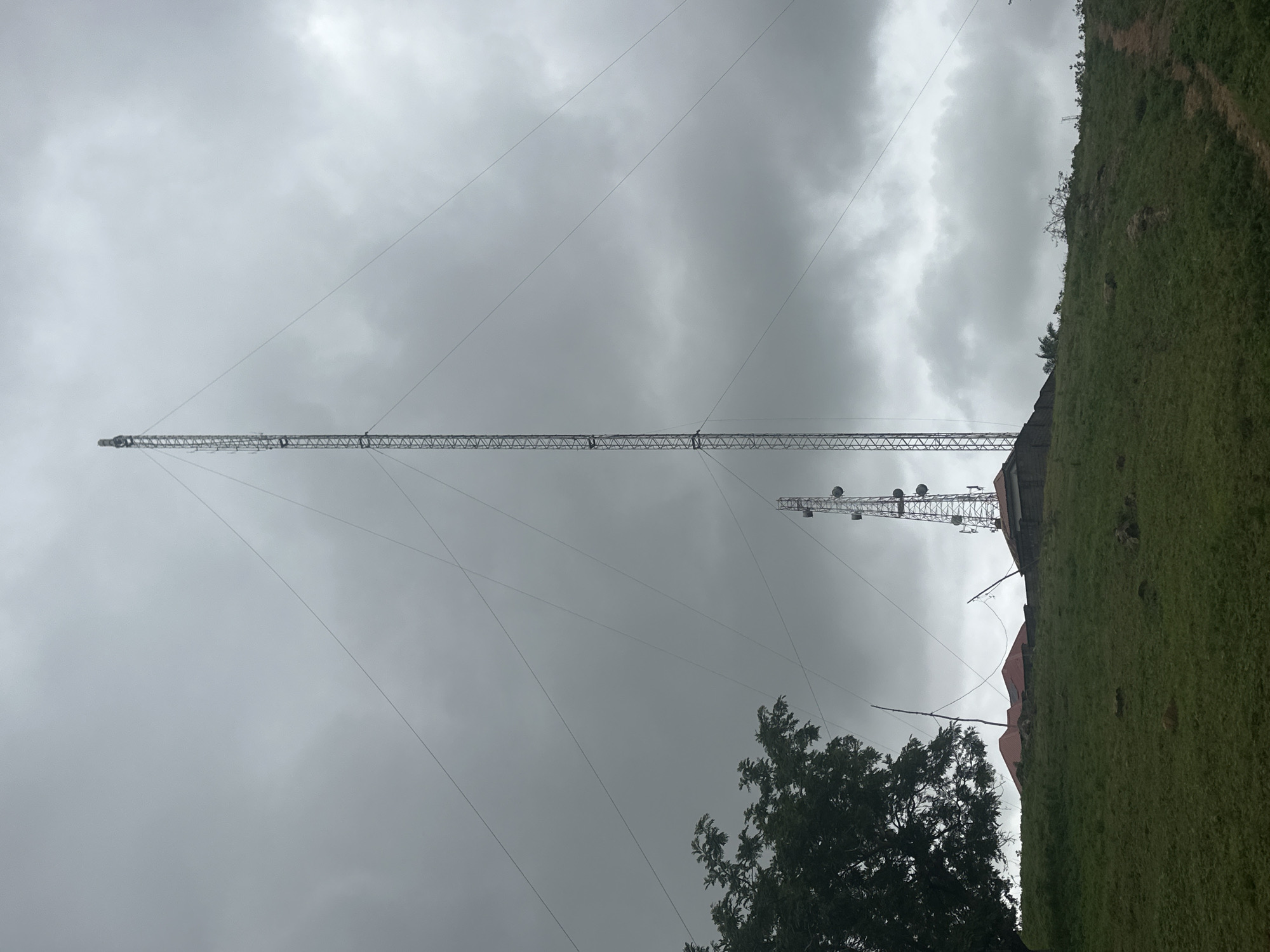 This image depicts an outdoor scene dominated by communication towers under a cloudy sky. The primary subject is a very tall, lattice-style tower, likely a radio or television transmission tower, stretching almost the entire height of the frame. Multiple guy wires extend from the tower to anchor points, creating a web-like pattern against the sky. In the mid-ground, a shorter, similar tower with antennas and dishes is visible, situated near a building with a reddish-brown roof. The background features a lush, green, grassy hillside with some trees. The weather appears overcast, with a heavy, grey cloud cover. The scene suggests a location with infrastructure for broadcasting or telecommunications, possibly in a somewhat rural or elevated area given the hillside. The orientation of the image, with the ground on the right, suggests it was taken with the camera rotated. There is no visible text.
