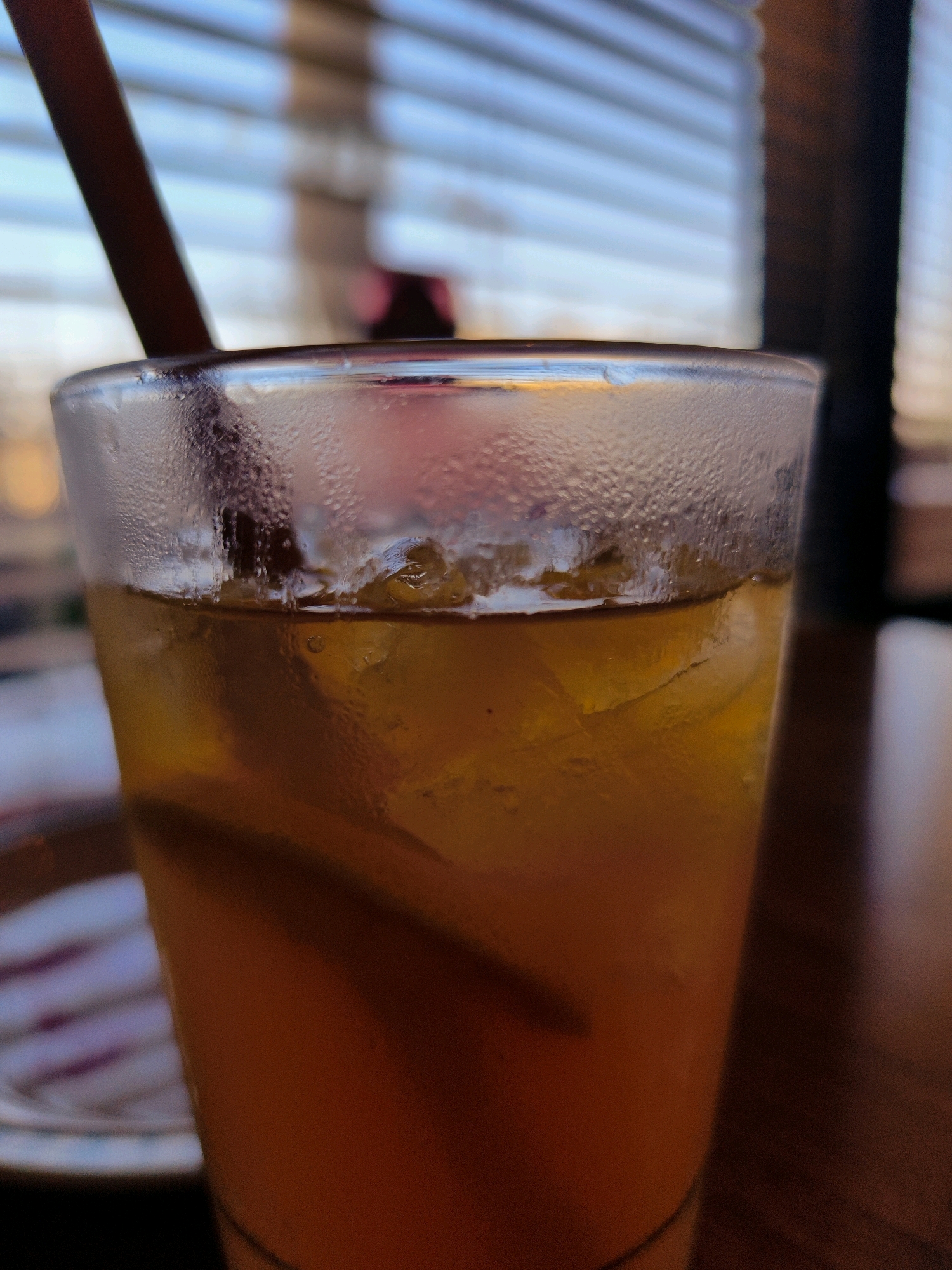 A close-up, low-angle shot focuses on a clear glass filled with iced tea and ice cubes. A dark-colored straw is visible emerging from the top of the glass. The exterior of the glass is covered in condensation. In the background, horizontal blinds are visible, with a soft focus on the outside environment, suggesting a setting by a window. The overall impression is of a refreshing beverage. The image was captured in Netivot, Israel, on the street called Ha'Nasi Re'uven Street.