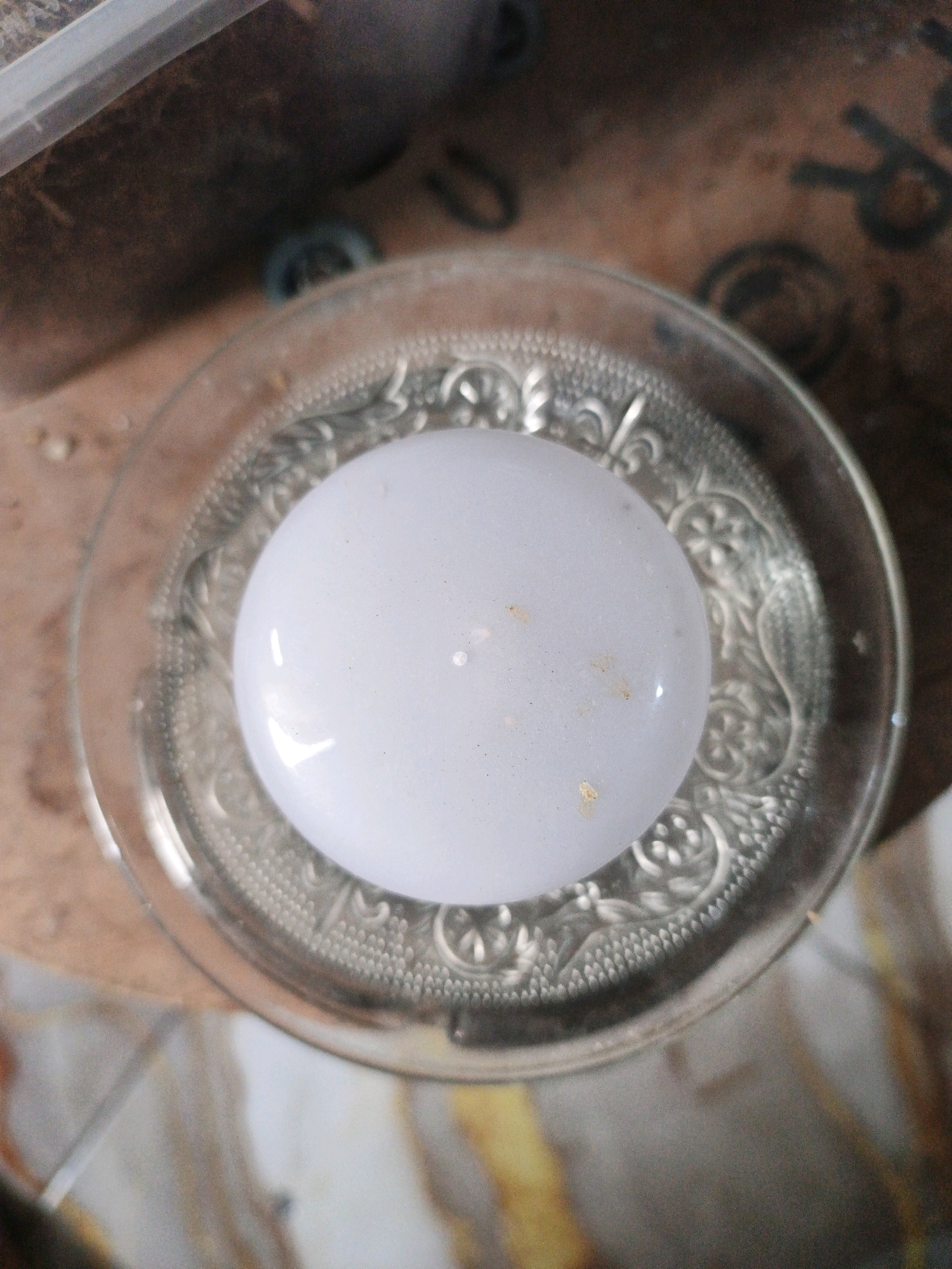 An overhead close-up shot shows a clear glass dish with an ornate rim, filled with water. A smooth, white, spherical object, possibly a ball or lightbulb, rests submerged in the water. The background is blurred, showing a wooden surface with some scattered debris and darker, indistinct objects. The scene is indoors and appears to be a still-life composition. There are no people or animals visible. The location of Ketegan, Indonesia, is not discernible from the visual information provided in the image.