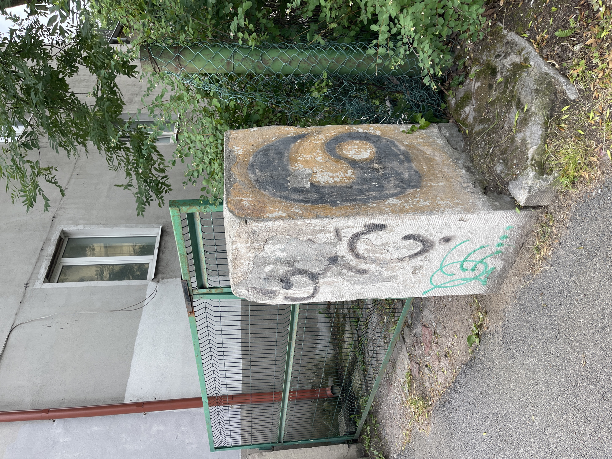 A rough-hewn stone pillar, approximately one meter tall, stands on a paved area next to a green metal fence. The pillar has graffiti on it, including a large black yin-yang symbol on one side and green squiggly lines on another. Behind the fence is a building with a window. To the right of the pillar and fence is dense green foliage and a chain-link fence. The scene appears to be in an urban environment in Kaliningrad, Russia.