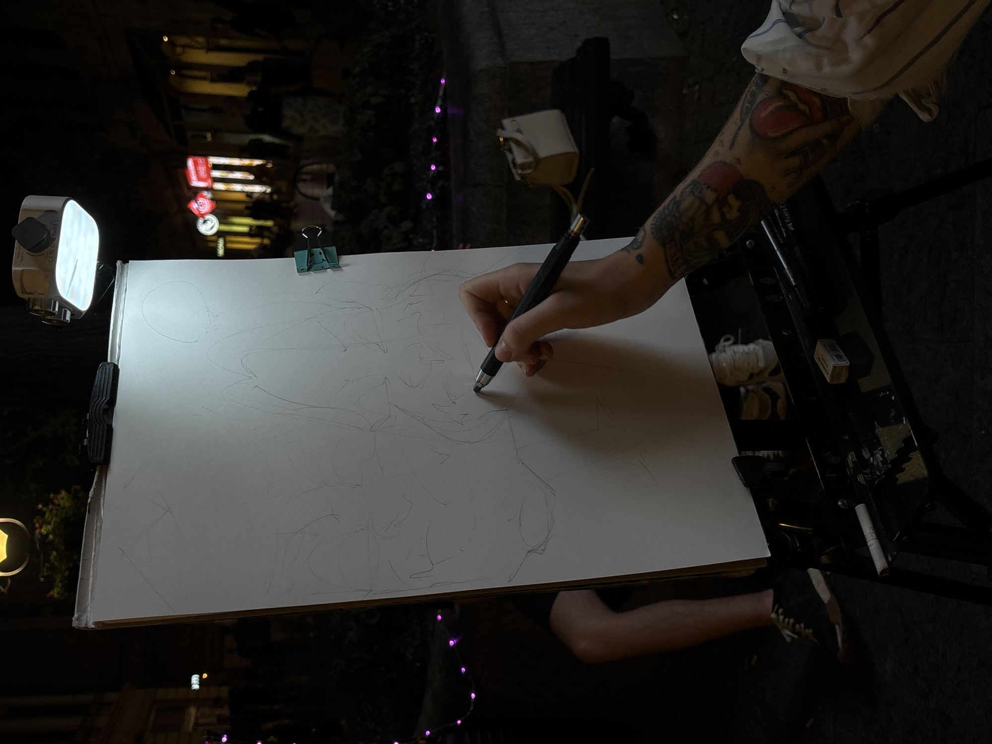 The image depicts an artist sketching on a canvas at night in an urban setting. The artist's hand, adorned with tattoos, holds a pen and is actively drawing on the canvas, which is illuminated by a bright LED light attached to the top of the easel. The background is dark, with blurred lights from storefronts suggesting a lively street scene. A person's legs and feet are visible to the left of the canvas, indicating they are sitting and possibly posing for the artist. A small white handbag rests on a dark surface behind the easel. The overall atmosphere is one of creative focus amidst the ambient city night. Without specific identifying markers, the exact location within Belgrade, Serbia cannot be definitively determined.