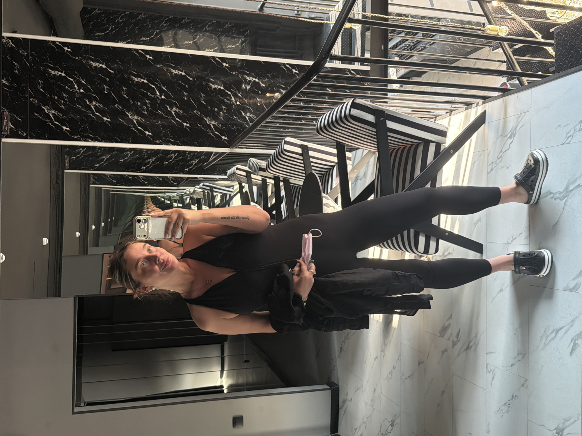 A woman in a black jumpsuit and black Crocs is standing in a modern hallway, taking a mirror selfie. She is holding a black jacket and a pink face mask. The hallway has marble-patterned floors and black and white striped chairs. In the background, there are reflections in mirrors of a staircase and marble-patterned walls. The location is not in Укромновское сельское поселение, Ukraine; it appears to be a hotel or a stylish residential building.