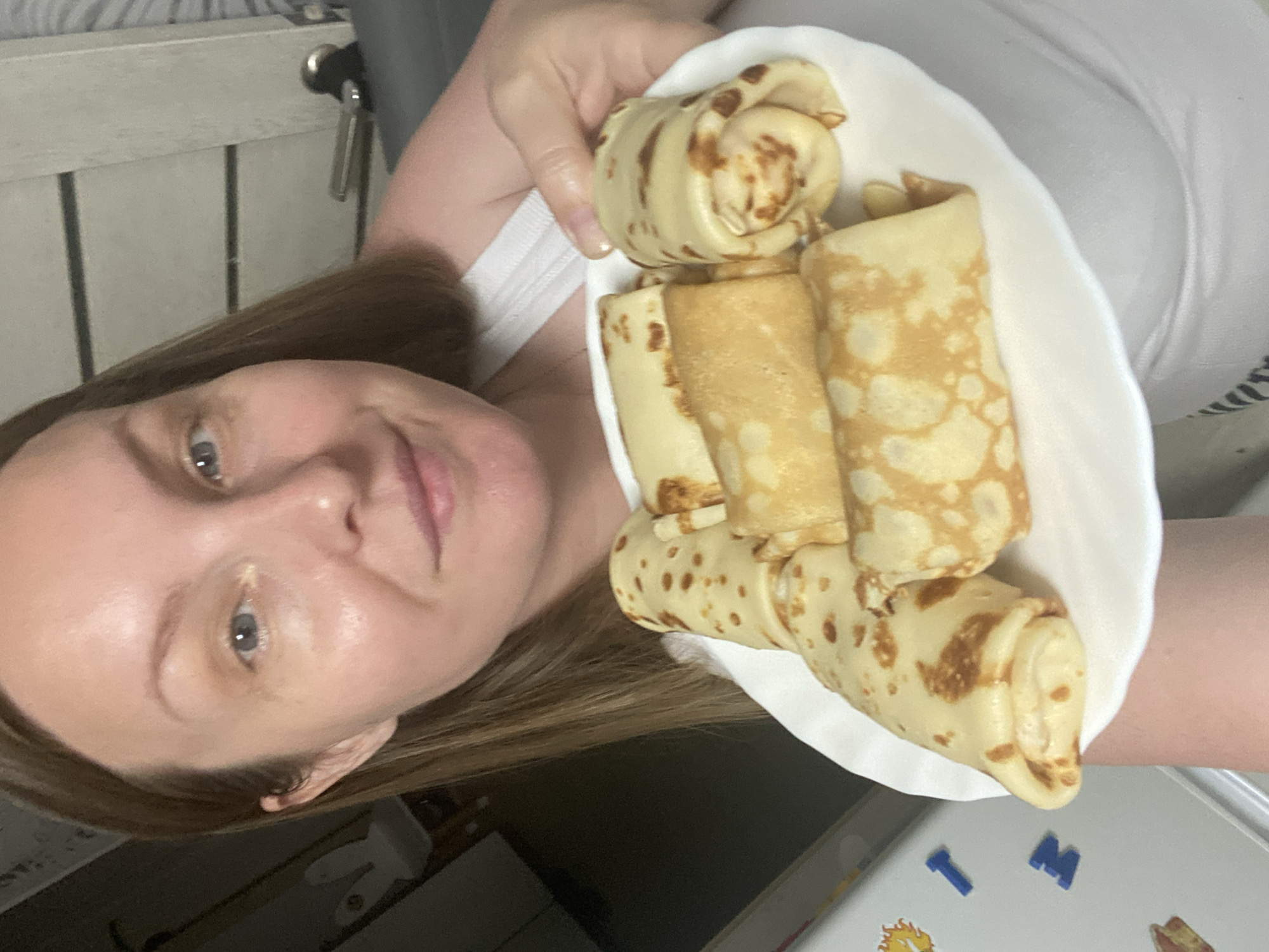 A woman is holding a white plate with several rolled crepes in front of her face. The crepes are light brown with darker brown spots and appear to be filled. The woman has light skin, brown hair, and blue eyes. She is wearing a white top. The background is out of focus, but a light-colored wooden cabinet and a refrigerator with magnets can be seen. The presence of magnets suggests a domestic kitchen setting, and the letters "T" and "M" are visible on the magnets, along with a sun drawing. The location Noviy, Russia, cannot be confirmed based on the image.