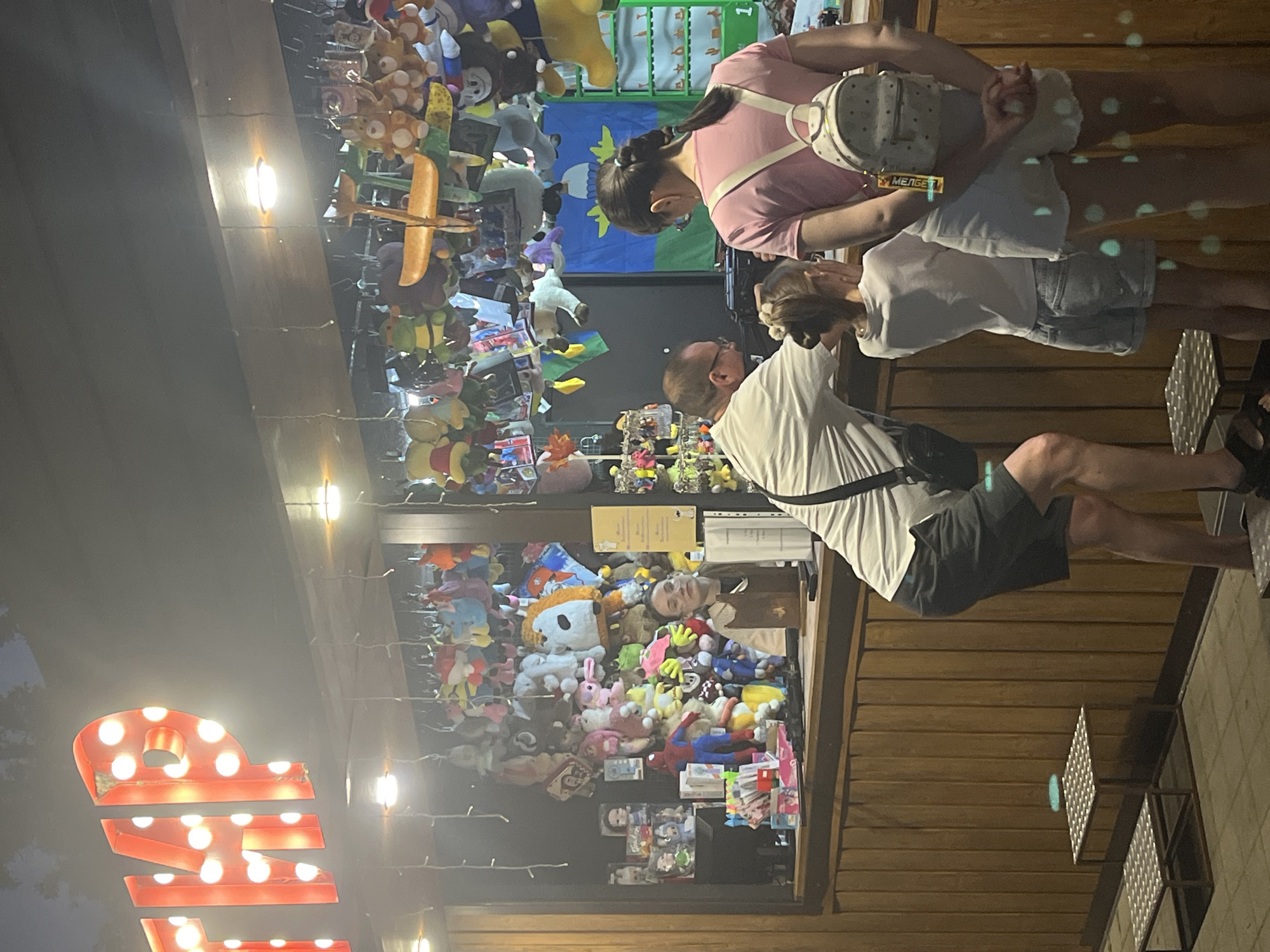 This image depicts an outdoor stall selling plush toys, likely in a recreational area. A red, illuminated sign spelling out "ТИР" (which translates to "Shooting Gallery" in Russian) is visible at the top of the stall. The stall itself is lined with wooden panels and is filled with numerous colorful plush toys hanging from the ceiling and displayed on shelves. A woman with dark hair, wearing glasses and a vest, is working behind the counter. In front of the counter, a man wearing a light-colored t-shirt and dark shorts is leaning over, seemingly interacting with a young girl who is wearing a white t-shirt and denim shorts. A young woman with dark hair in a braid, wearing a pink t-shirt and white shorts, stands beside the girl, with a small backpack on her back. Two small metal step stools with perforated tops are placed on the ground in front of the stall. The lighting suggests it is evening or nighttime. The scene is likely in Krasnodar, Russia, given the presence of a Cyrillic sign.