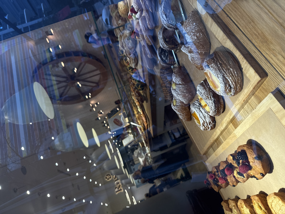 The image displays the interior of a Rosetta Bakery establishment located in Miami, United States of America, viewed through a reflective glass storefront.
In the foreground, an assortment of pastries is arranged on light wooden display boards. These include several croissants heavily dusted with powdered sugar; some are plain, while others feature fillings such as peach or apricot slices, or a creamy white substance. Darker, layered pastries, possibly Kouign-amann, are also visible, along with individual golden-brown muffins or small cakes topped with red raspberries, dark blackberries, and blueberries.
The bakery's interior, visible through the reflective glass, features a modern, brightly lit environment. A large, decorative wooden wheel, resembling an old mill wheel, is prominently mounted on a textured white wall in the midground. Display cases line the counters, showcasing various breads and additional baked goods. The "Ros" segment of the Rosetta Bakery logo is visible on an interior wall in the upper left. Several individuals, appearing as both patrons and staff, are present within the bakery, their forms somewhat blurred due to the viewing angle and reflections. No specific actions or interactions are clearly discernible among them. Reflections on the glass surface show faint blue hues and vertical lines, indicating elements of the external urban environment.