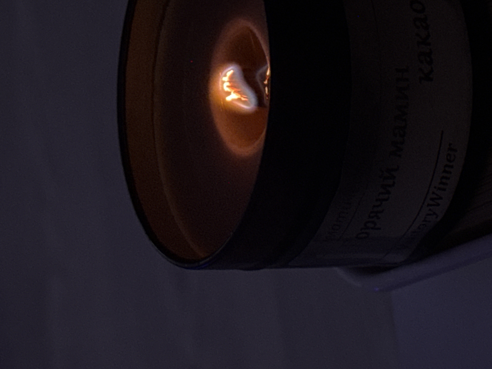 A lit candle, partially obscured by a dark label with Cyrillic text, sits in an ambiguous setting. The flame is visible within the wax pool. The location of Ново-Никольское, Russia is not visibly discernible from the provided image.