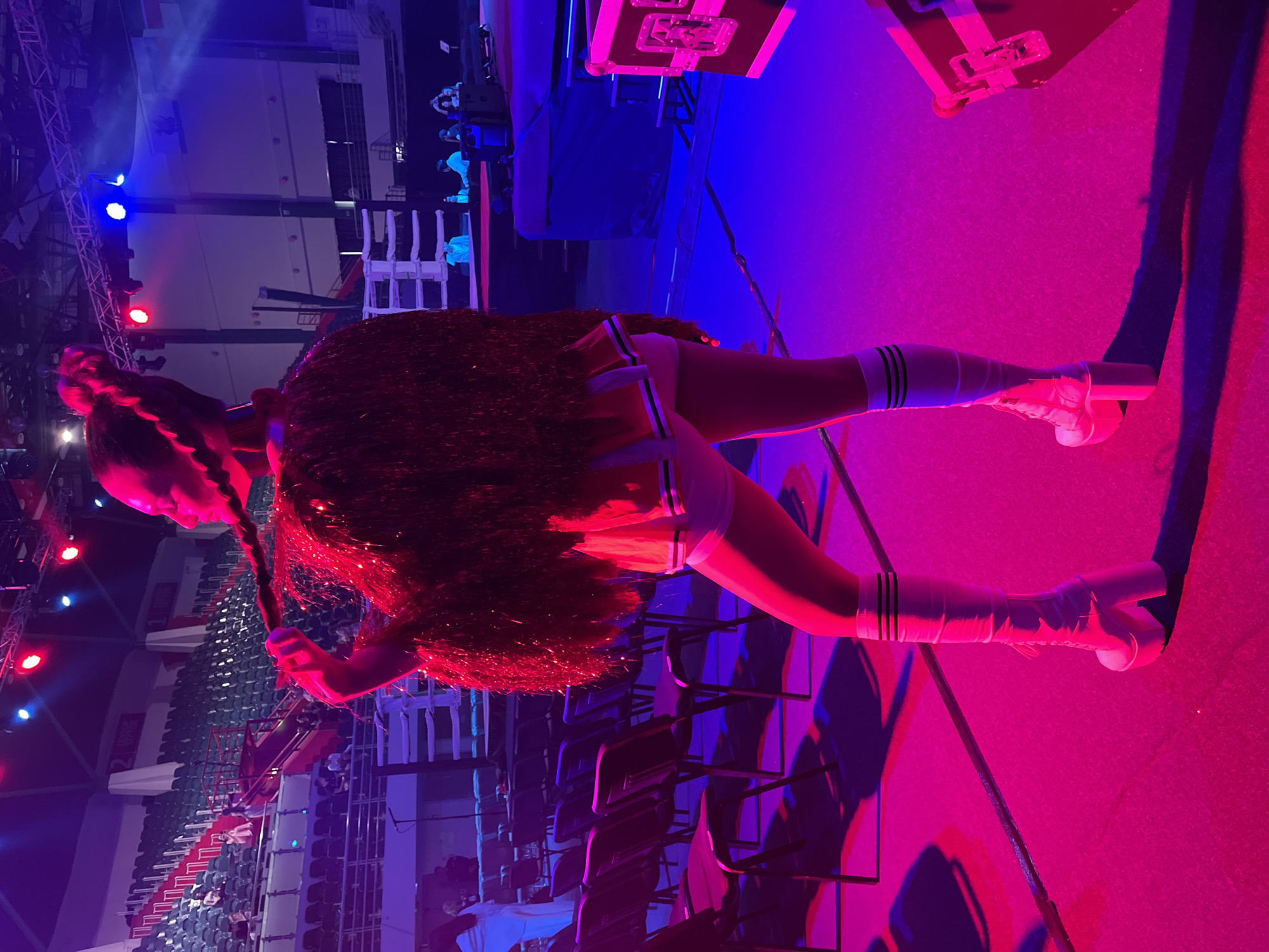The image shows a woman, likely a performer or entertainer, holding what appears to be a microphone, standing in an indoor arena or event space. She is dressed in a costume consisting of a short skirt and boots with striped knee-high socks. She has long hair in a braided style. The background includes rows of empty seats, stage lighting, and equipment. The scene is bathed in blue and pink lighting. The location is possibly in Kazan, Russia.