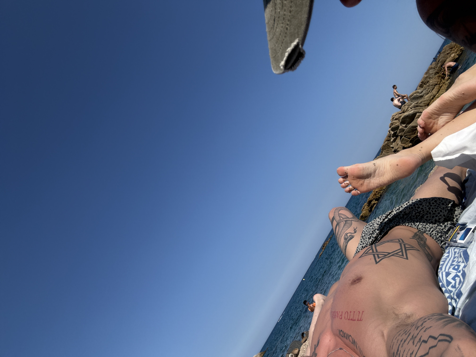 In Castell d'Aro, Platja d'Aro i s'Agaró, Spain, a person lies on a towel on a beach, with a clear blue sky overhead and the sea in the background. The person is partially visible, showing tattoos on their torso and legs, including a Star of David and a snake. They are wearing patterned swim trunks. Another person's feet are visible in the foreground, with a cigarette pack placed near them. In the sea, two individuals are seen near rocks, with one appearing to be jumping into the water. The overall scene depicts a sunny day at the beach with people enjoying the water and sun.