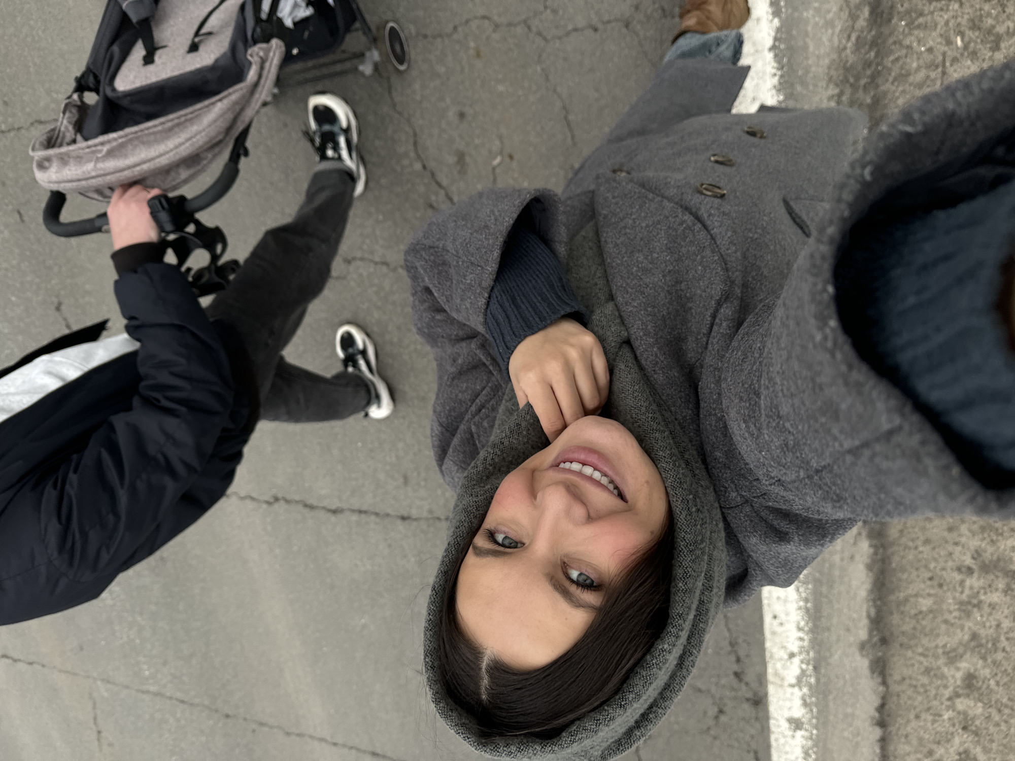 A woman in a gray hooded coat smiles upward into a selfie shot. Next to her is a person wearing dark pants, black and white sneakers, and a dark jacket, pushing a stroller. They are standing on a gray surface resembling concrete or asphalt. Patches of what appears to be salt or snow are visible on the right edge of the frame. The precise location in Красный Маяк, Russia is not discernible from the provided image.