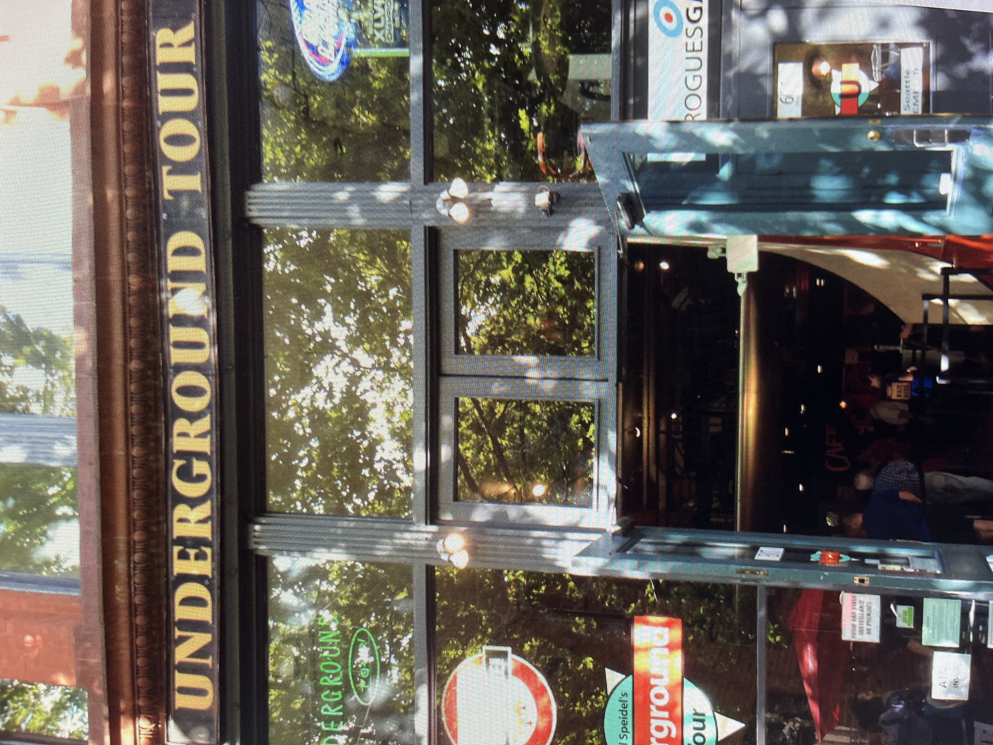 This media file shows the storefront of the "Underground Tour" business. The facade features large windows with reflections of trees and sunlight, suggesting an outdoor setting during the daytime. A prominent vertical sign on the left side of the image reads "UNDERGROUND TOUR" in large yellow letters. Through the windows, there are glimpses of the interior, including the words "CAFE" and various signs and posters. One sign, partially visible, mentions "ROGUESG" and another has the number "60" and "Seattle EMETE". There are also what appear to be people inside the building, though their features are not clearly discernible due to the reflections and lighting. The overall impression is a busy commercial establishment offering tours of an underground area. The scene suggests a bright day, and the presence of a tour business implies a tourist-oriented location. The "Speidel's rground our" text indicates part of the business's name or service offering.