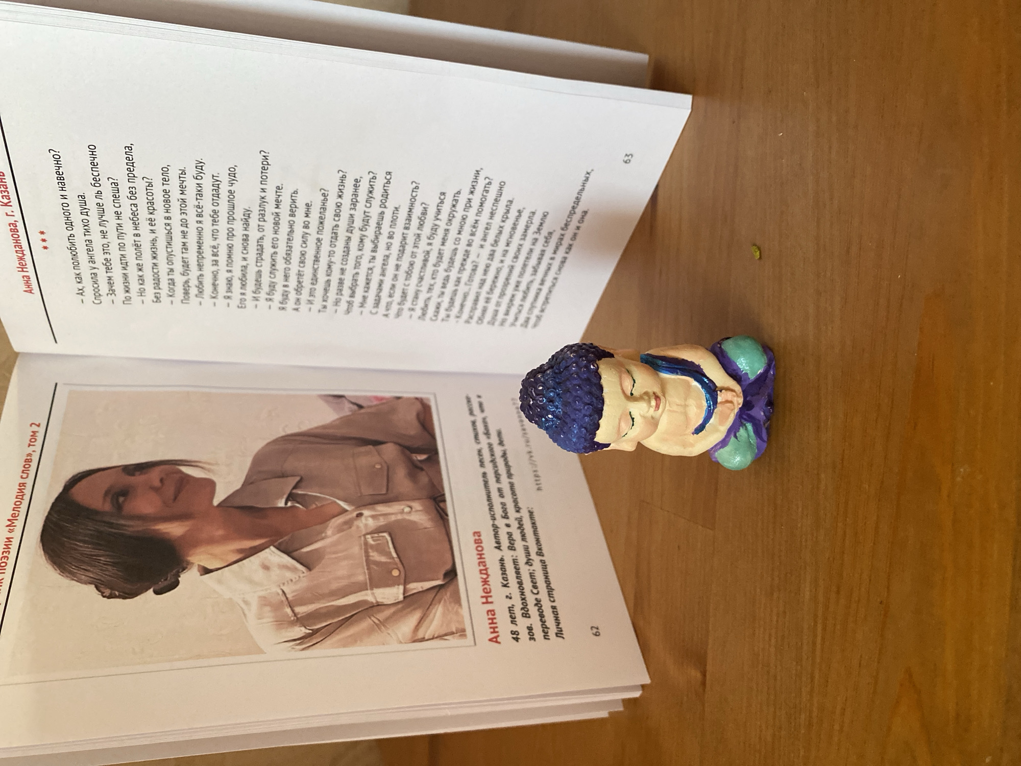 The image shows an open book with a portrait of a woman on the left page and a poem on the right page. The woman in the portrait is looking to the right and smiling. She has dark hair pulled back in a bun and is wearing a light-colored collared shirt. Below the portrait, text identifies her as "Anna Nezhdanova" and provides biographical details, including her age (48), location (Kazan), and her role as a singer, songwriter, and performer. Her inspirations are listed as "faith in God, verses of Russian poetry, music, and love for the world; the souls of people, the beauty of nature." A personal social media link is also provided.
The right page features a poem titled "Melody of words, tome 2" with the author's name "Anna Nezhdanova, g. Kazan" at the top. The poem is written in Russian.
In the foreground of the image, a small, colorful statue of a meditating Buddha is placed on a wooden surface. The wooden surface has a visible grain pattern. The overall lighting suggests an indoor environment. There are no visual elements in the image that indicate the city of Zelenodol'sk, Russia.