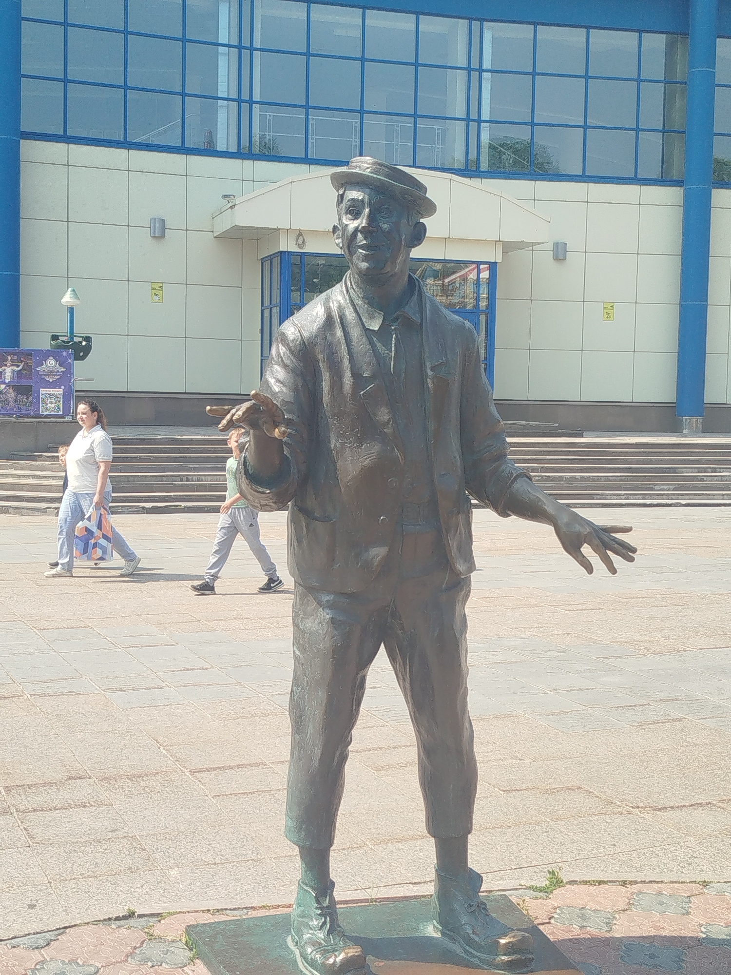 A bronze statue of a man wearing a hat, jacket, and trousers stands in an outdoor plaza in Tyumen, Russia. The statue is life-sized and positioned in the foreground, facing slightly to the right. The man's right hand is extended forward, and his left hand is slightly bent at the elbow. In the background, a modern building with large blue-tinted windows and light-colored cladding is visible. Two people are walking in the plaza: a woman in a white shirt and light-colored jeans carrying a patterned shopping bag, and a child in a light-colored shirt and grey trousers walking behind her. The plaza pavement is made of light-colored square tiles.