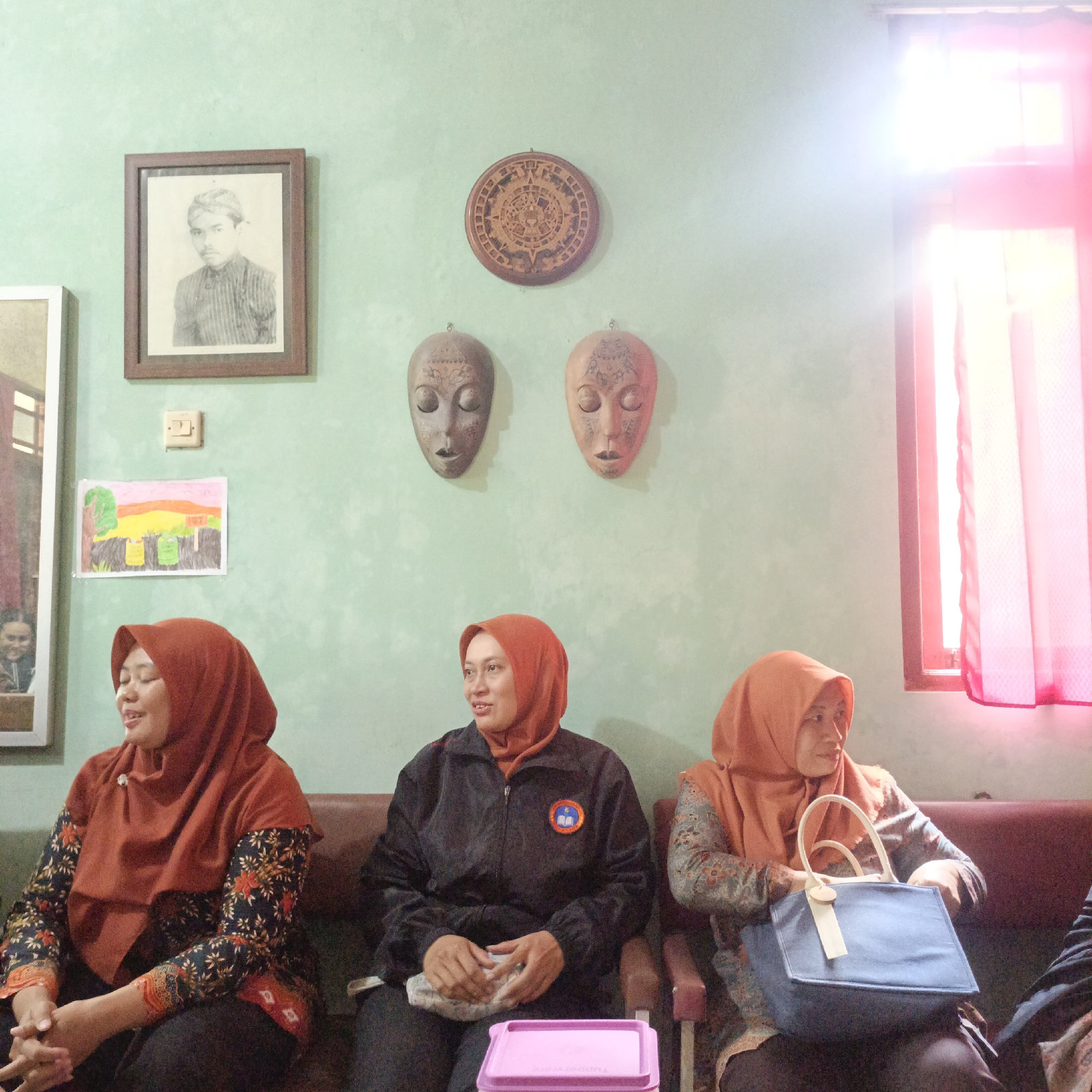Three women are seated on a sofa against a light green wall. The woman on the left wears a rust-colored hijab and a dark patterned long-sleeved top. She is looking to her left. The woman in the middle wears a rust-colored hijab and a black jacket with a circular logo on the left chest. She is holding a light-colored object in her lap and looking forward. The woman on the right wears a rust-colored hijab and a patterned long-sleeved top. She is holding a blue tote bag with a light-colored tag in her lap and looking to her right. On the wall above the women are several decorative items: a framed black and white portrait of a man, a round carved wooden disc, and two carved masks. To the left of the portrait is a light switch. Below the portrait and to the left of the women is a framed child's drawing of trees, a landscape, and two bins. To the right of the women is a window with pink curtains and bright light shining through. The overall environment appears to be indoors, possibly a home or community space, and the scene is consistent with a social gathering or meeting in Salatiga, Indonesia.