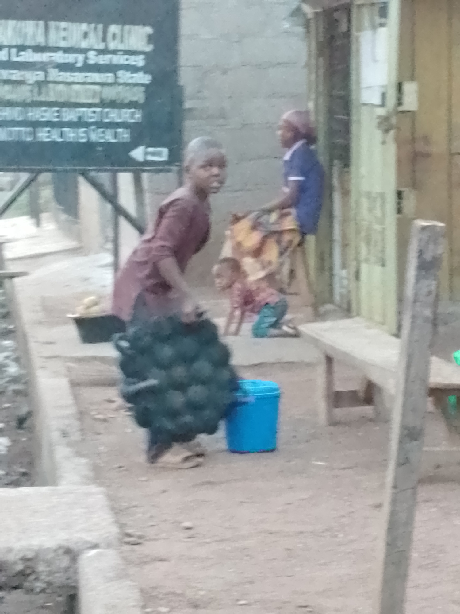 The image shows a street scene in Akwanga, Nigeria, as identified by the sign reading "AKWANA MEDICAL CLINIC" and "KWARANYA NASARAWA STATE." A child is carrying a large, heavy object made of black circular elements, appearing to be either a heavy metal sculpture or a device. Next to the child is a blue bucket. In the background, a woman wearing a blue shirt sits on a stool, and another child is kneeling on the ground. A sign indicating "SAND HASKE BAPTIST CHURCH" and "MOTTO HEALTH IS WEALTH" is also visible. A simple wooden bench is positioned near a building's entrance.