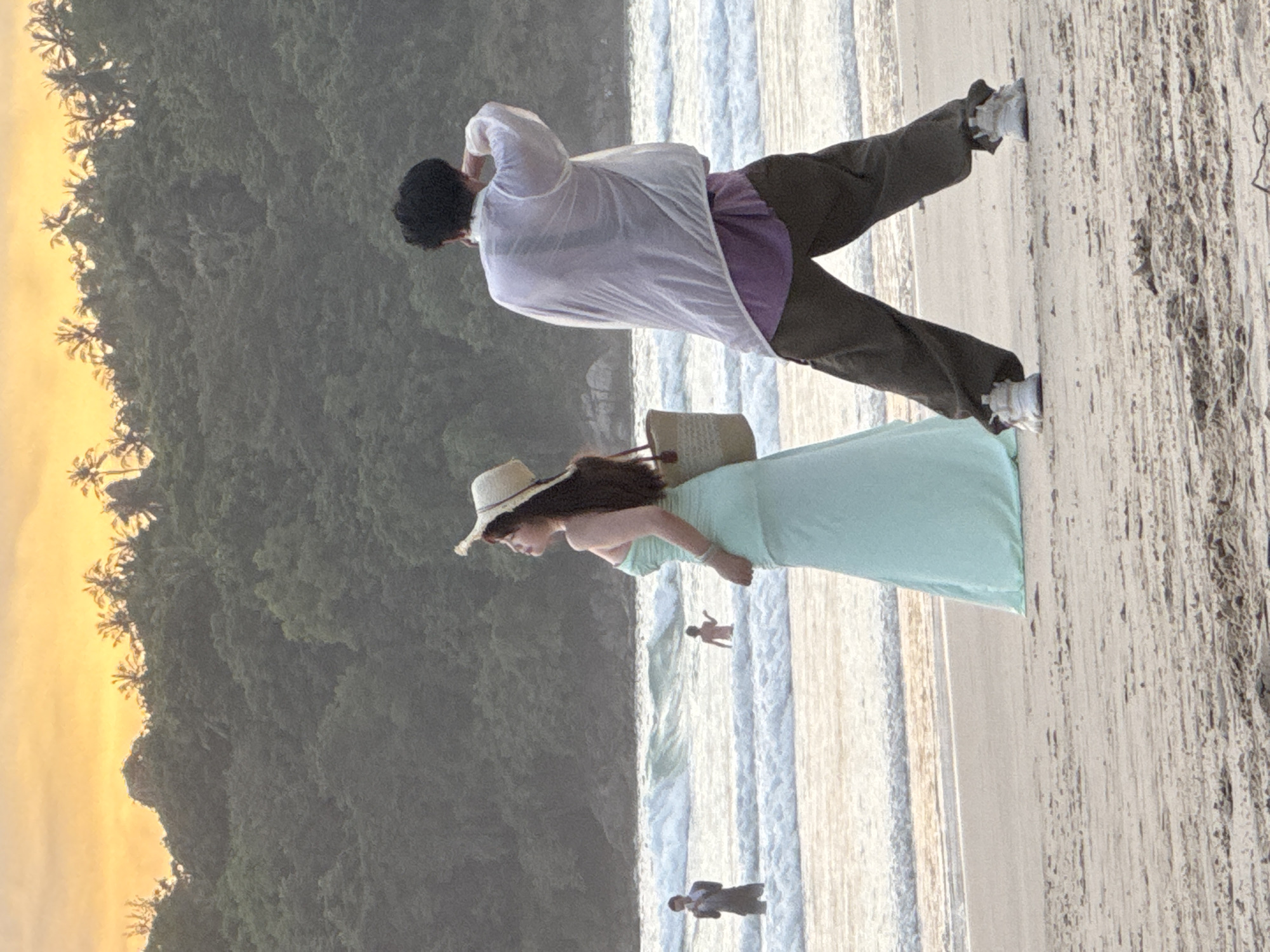 In Karon, Thailand, two individuals stand on a sandy beach facing the ocean as the sun sets. In the foreground, a woman with long, dark hair wears a strapless mint-green dress and a straw hat. She carries a woven tote bag over her right shoulder. To her right, a person with dark hair wears a translucent white shirt over a purple t-shirt and dark pants. They are turned away from the camera, with their right arm raised to their head. In the background, two other individuals are visible in the water, with waves crashing around them. The ocean stretches out to the horizon, meeting a sky painted with hues of orange and yellow from the setting sun. Lush green hills, dotted with palm trees, form a backdrop to the scene.