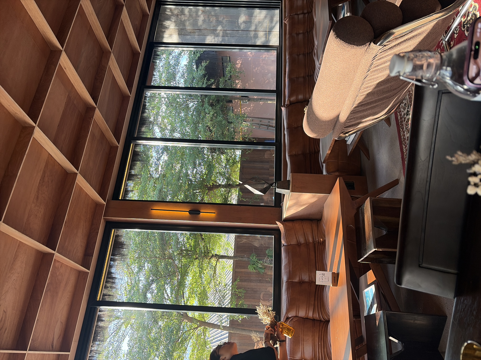 An indoor seating area in a cafe in North Kuta, Indonesia, features a wooden coffered ceiling, large windows overlooking lush trees and a wooden fence, and brown leather sectional sofas. A long, low wooden table is situated in front of the sofas, with small wooden stools and a dark wooden table in the foreground. A single person, partially visible on the left, is holding a small orange vase with dried flowers. An abstract sculpture stands on a wooden pedestal near the window. A bottle of water with a silver stopper is in the foreground. The lighting suggests late afternoon sun.