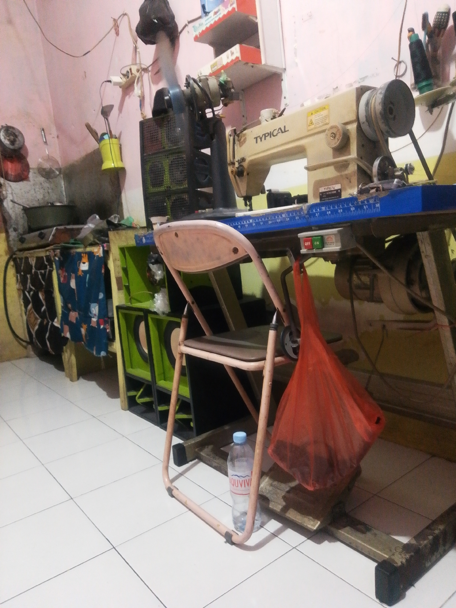 The image depicts an indoor scene, likely a workshop or sewing area, with a pink-painted wall and tiled floor. A "TYPICAL" brand sewing machine is prominently displayed on a table with a blue ruler attached to the front edge. To the left of the sewing machine, a black fan with a metal cage sits on top of a green cabinet that appears to house speakers. Various household items and tools are visible, including a yellow bucket with kitchen utensils, a dark-colored bag hanging from the table, and a bottle of water on the floor. The overall impression is one of a cluttered, functional workspace. The location is identified as Bohar, Indonesia.