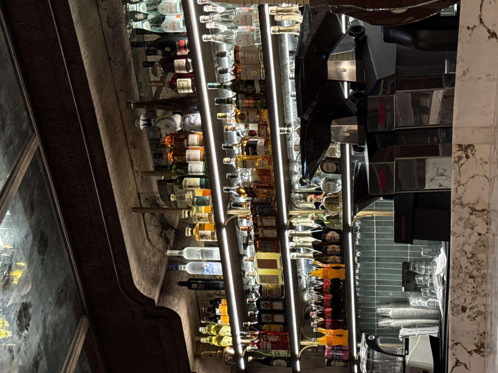 The image depicts the interior of a bar, likely upscale, stocked with numerous bottles of alcohol arranged on illuminated shelves. The variety of bottles suggests a full bar, capable of serving a wide range of drinks. To the right of the bar, a beverage dispensing system and clean glassware indicate an area for drink preparation and service. The architectural style of the bar includes dark wood detailing and what appears to be decorative stone or marble, implying a sophisticated and perhaps historical location. The specific location in Cape Town, South Africa, is not discernible from the image alone, but the overall aesthetic suggests a venue that caters to a discerning clientele.