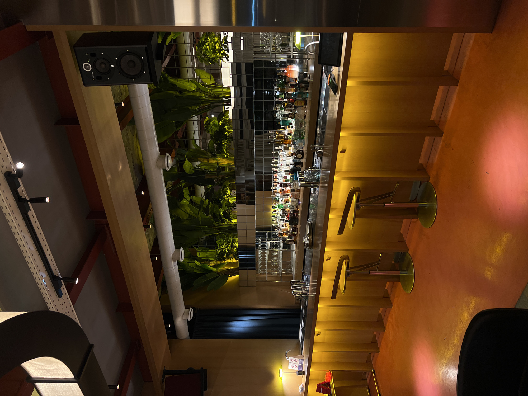 The image depicts the interior of a modern bar, likely located in Yerevan, Armenia. The bar features a long, illuminated counter made of golden wood, with several bar stools with yellow seats and dark backs arranged in front of it. Behind the counter, a mirrored backsplash displays an array of liquor bottles and glassware, suggesting it is fully stocked and operational. Lush green plants are strategically placed behind the bar, adding a touch of nature to the ambiance. Overhead, industrial-style lighting fixtures and exposed pipes contribute to the contemporary aesthetic. A black speaker is mounted on the wall above the plants. The floor is a vibrant orange-red, reflecting the warm lighting of the bar. The overall impression is one of a stylish and well-equipped establishment, ready to serve patrons.