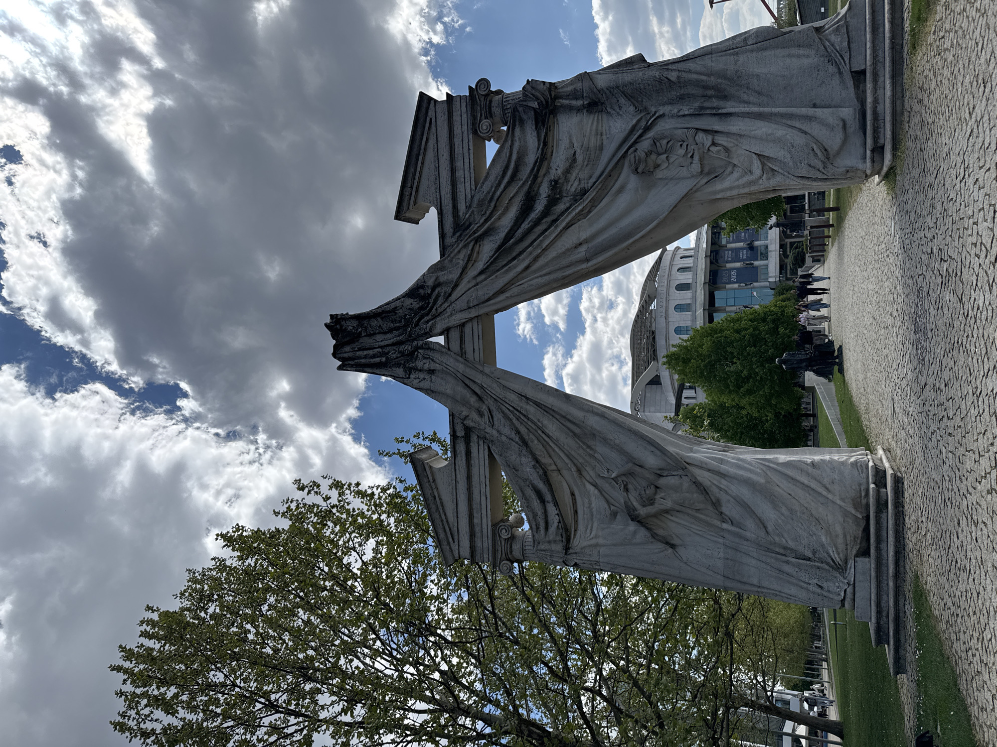 This image captures the Memorial to the Victims of the German Occupation, located in Budapest, Hungary. The memorial consists of two abstract, draped stone figures representing the Archangel Gabriel, positioned opposite each other to symbolize the division and suffering caused by the Nazi occupation during World War II. The figures stand at the entrance to a walkway leading towards a larger building complex that can be seen in the background. In the distance, people are walking along the walkway near the building. The sky above is filled with dynamic clouds, ranging from bright white to dark gray. Below, trees with green leaves frame the lower portion of the composition. The foreground features a paved path with stones.