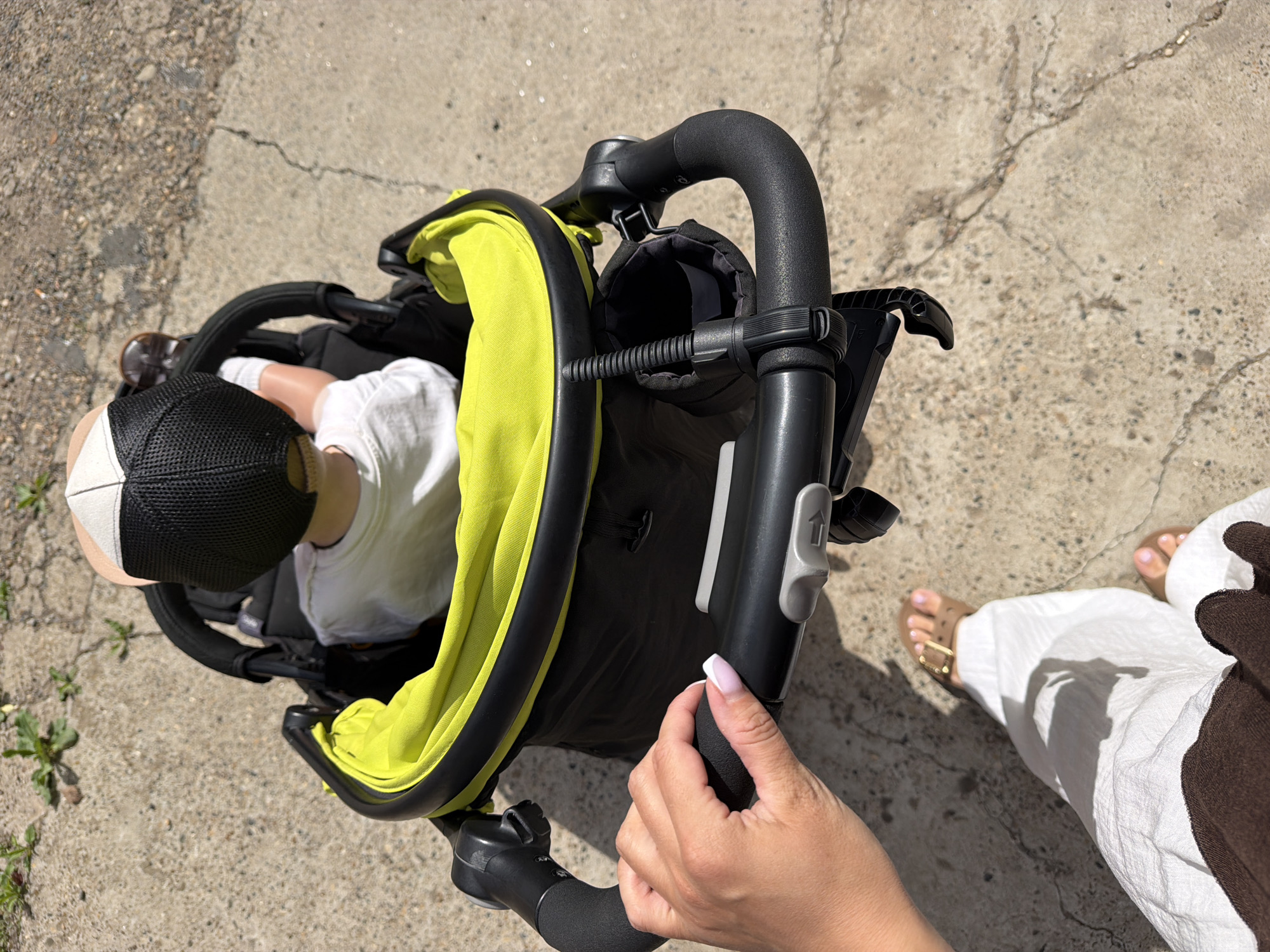 The image shows a close-up, high-angle view of a person's hand gripping the handle of a stroller. The stroller has a black frame with a bright yellow interior lining. A child, wearing a white shirt and a black and tan cap, is seated in the stroller, facing away from the camera. The stroller is on a concrete surface, possibly a sidewalk or pathway, with some sparse vegetation visible in the upper left corner. The lower part of the image shows the legs and feet of the person pushing the stroller. They are wearing white pants and brown sandals. The scene appears to be outdoors on a sunny day, with shadows cast on the ground. There is no information in the image to suggest it is located in Votkinsk, Russia.