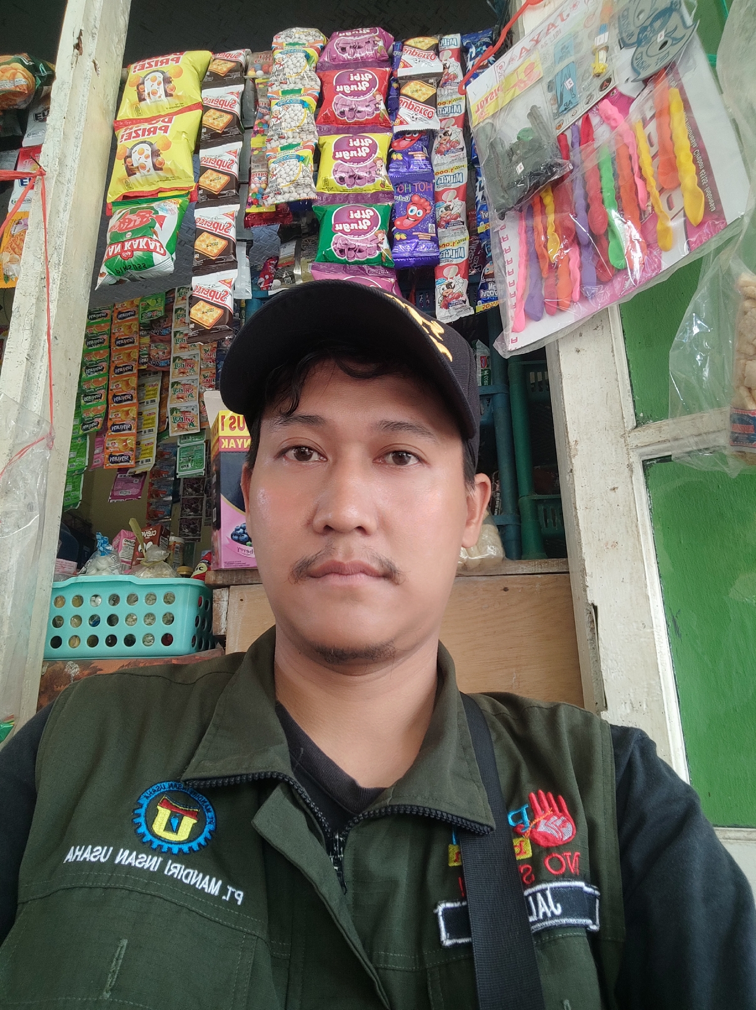 A man wearing a dark green vest with patches and a black baseball cap is taking a selfie in what appears to be a small roadside shop in Karangwangun, Indonesia. The shop is filled with various packaged snacks and small items displayed on shelves behind him. A turquoise plastic basket is visible to his left, containing more items. The man has a mustache and a short beard. The overall setting suggests a humble commercial establishment.