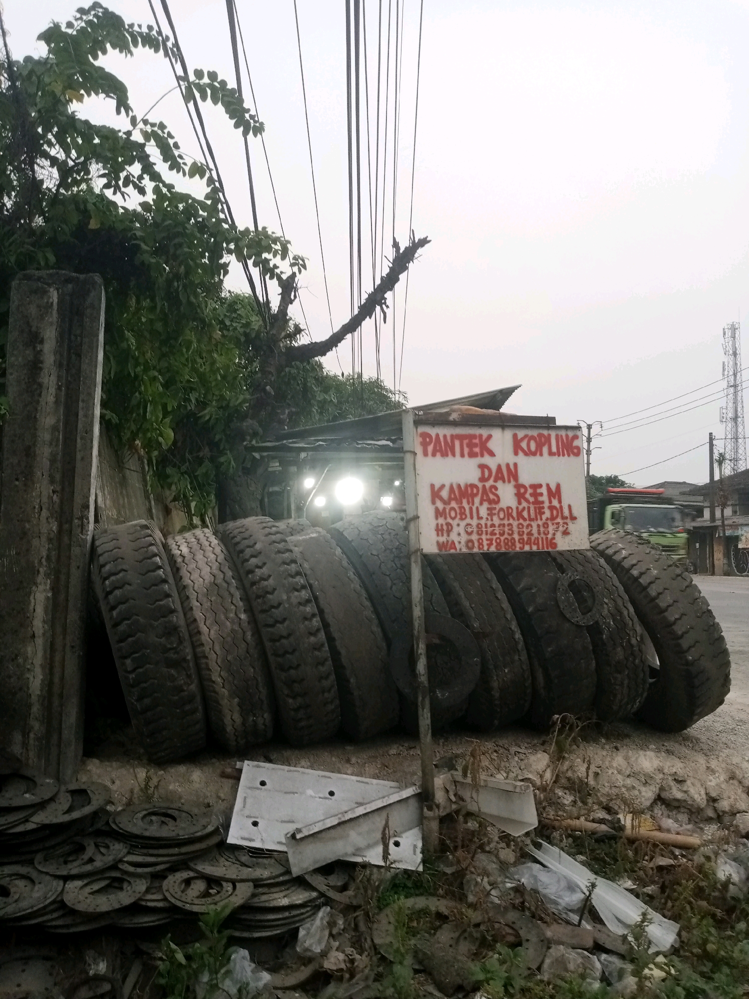 A close-up, low-angle shot captures a collection of used tires and brake discs piled on the side of a road in Balaraja, Indonesia. In the background, a sign with red lettering reads "PANTEK KOPLING DAN KAMPAS REM MOBIL FORKLIFT.DLL" and displays phone numbers. The sign is propped up by a metal pole, with a row of large tires stacked behind it. A concrete pillar stands to the left of the tires, partially obscuring a grey wall. The scene is framed by overhanging tree branches and a dense canopy of leaves. The sky is overcast, and the overall lighting suggests it is either early morning or late afternoon. The road surface appears to be dirt and gravel. In the distance, a green truck can be seen driving past, and other buildings and a telecommunications tower are visible. The ground in the foreground is littered with various debris, including more brake discs and discarded materials.