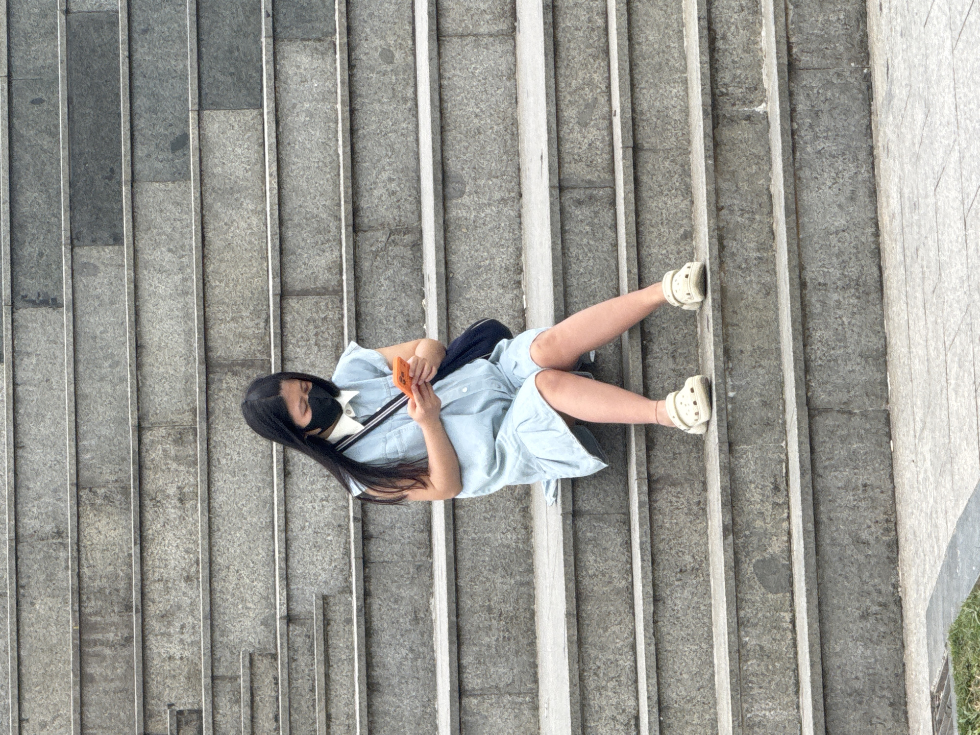 A young East Asian female wearing a light blue collared dress, a black face mask, and white clogs sits on a concrete staircase, looking down at her orange smartphone. She has long, dark hair and is holding the phone in her hands. A dark blue bag with a white strap is slung over her shoulder. The staircase appears to be made of grey stone with visible texture. The background is also composed of similar concrete stairs, creating a sense of depth. The lighting suggests an overcast day. The scene is set outdoors, likely in a public area with stairs, such as a park or a monument in Da Lat, Vietnam.