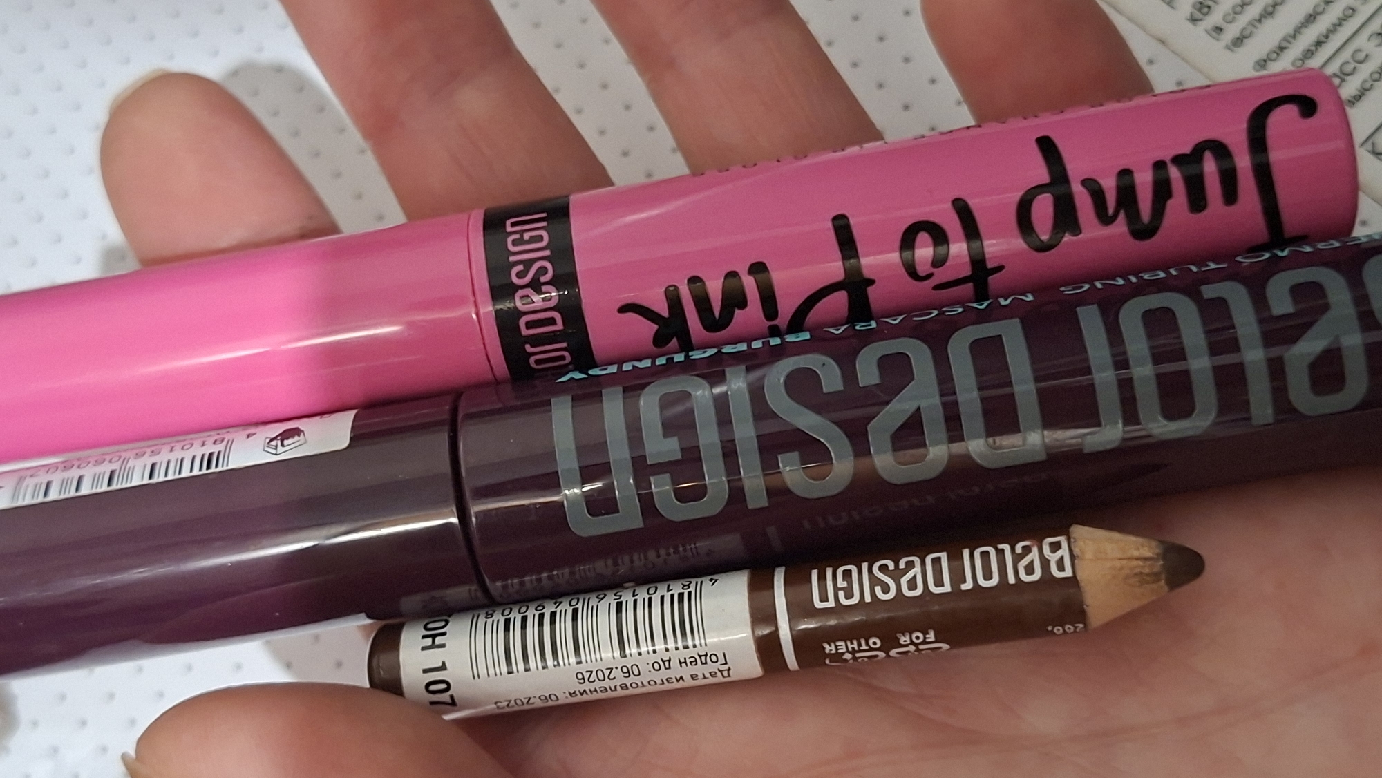 A person's hand holds three cosmetic products, a brown eyeliner pencil and two mascara tubes, one dark purple and one bright pink. The eyeliner pencil has "Belor Design" and "for other" written on it. The dark purple mascara tube has "Belor Design" printed vertically in large silver letters. The bright pink mascara tube has "OR DESIGN" and "Jump to Pink" printed on it, with "BURGUNDY" written vertically in smaller text. The background is a white surface with a subtle pattern of small gray dots. The products appear to be new, with the eyeliner pencil sharpened. The image is taken at a close-up, slightly angled perspective. There is no information or visual cues in the image that indicate the location in Забалоцкі сельскі Савет, Belarus.