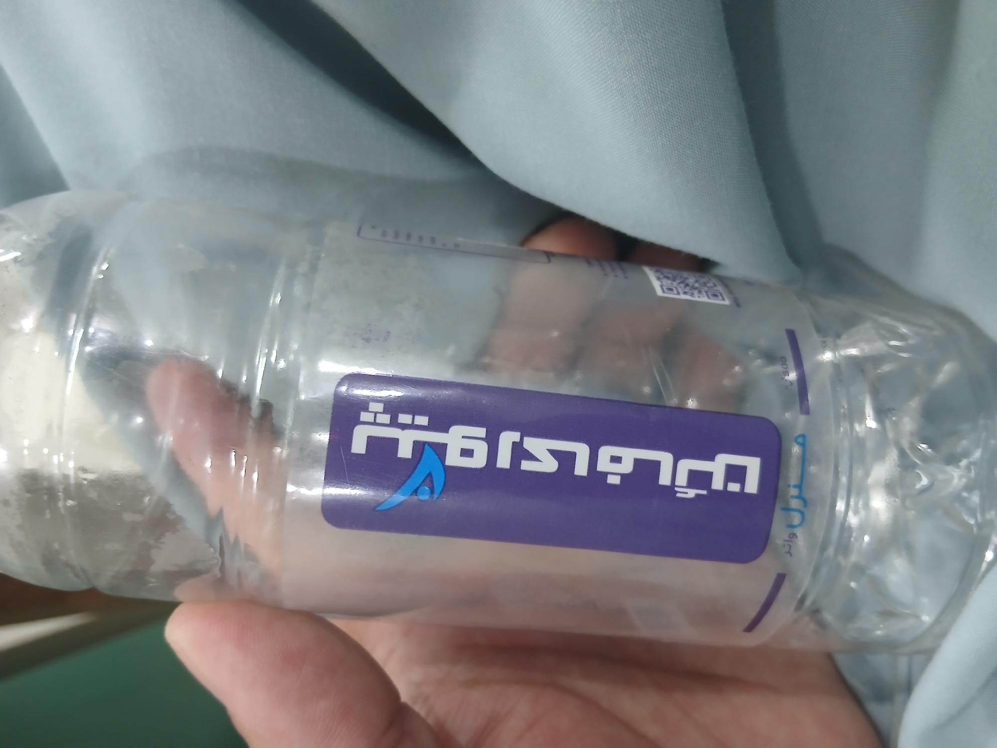 A clear plastic water bottle, held by a person's hand, is shown against a light blue fabric background. A purple label on the bottle features white Arabic script and a blue and white logo. Below the label, the word "MINERAL WATER" is visible in light blue text. A QR code is also present on the lower right side of the label. The scene appears to be indoors.