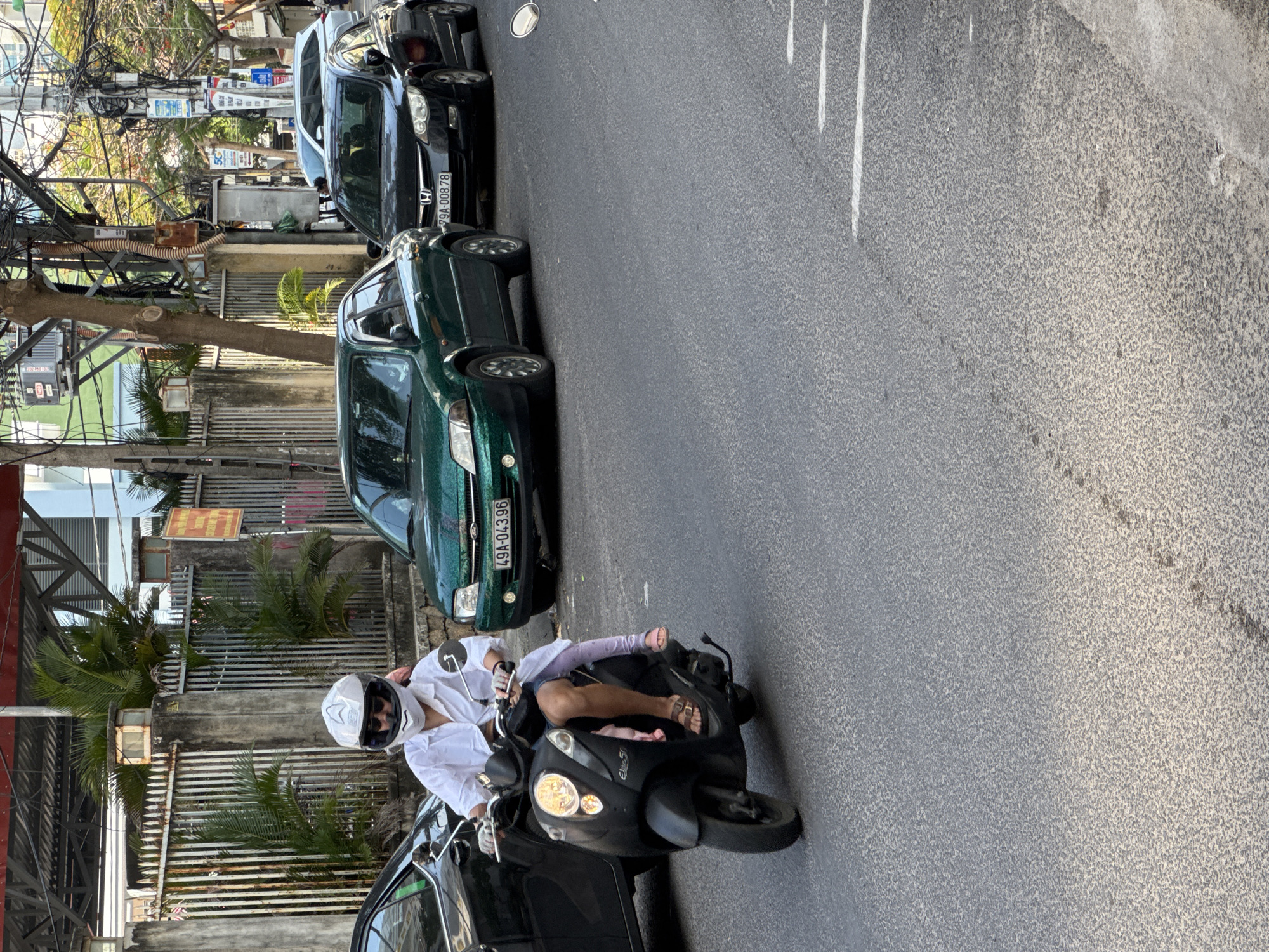 A person wearing a white long-sleeved shirt, white helmet, sunglasses, and dark shorts rides a black scooter. A child in a pink top and light purple leggings is seated in front of the rider. The scooter has a white logo on the front. The rider wears brown sandals, and the child wears pink sandals. The scooter has a white headlight and turn signals. The scene appears to be a street in Nha Trang, Vietnam, with traffic. A dark car is to the left of the scooter, and two cars, one green and one black, are behind the scooter. The green car has a license plate that reads "49A-043.96". The black car has a license plate that reads "79A-008.78". The background shows a fence, palm trees, and power lines.