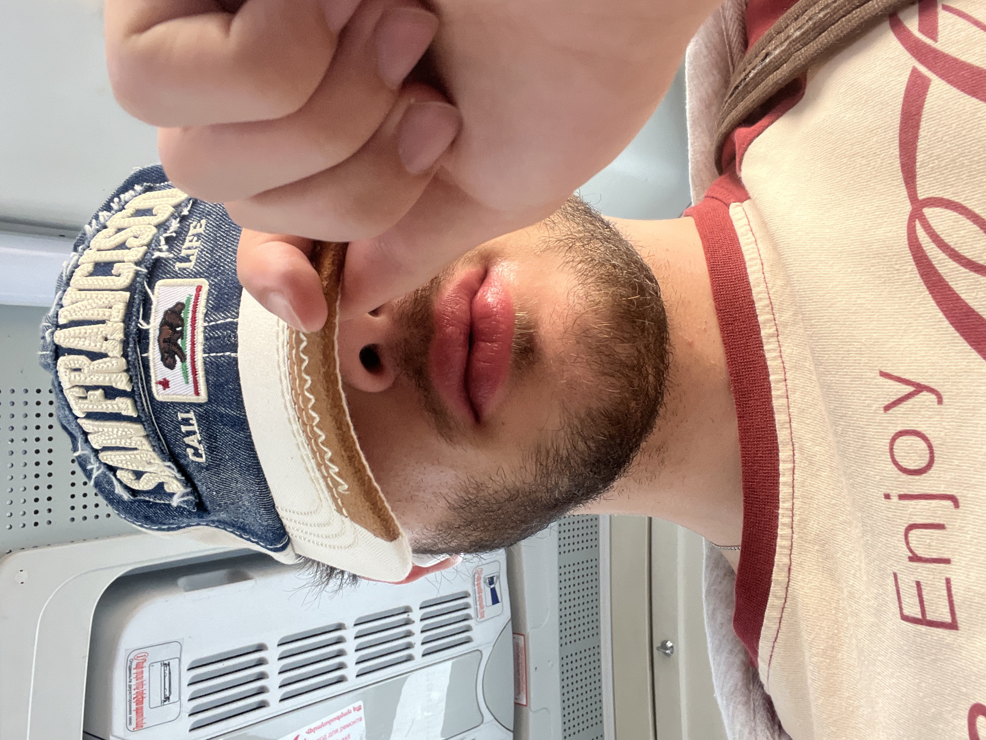 The image shows a selfie taken by a person wearing a "CALIFORNIA" baseball cap and a shirt with the word "Enjoy" on it. The person's face, partially obscured by a hand, is visible, revealing a beard. Behind the person is what appears to be an airplane interior, including an air conditioning unit with Armenian text. Given the presence of Armenian text, the location could potentially be in Yerevan, Armenia; however, this cannot be definitively confirmed from the image.
