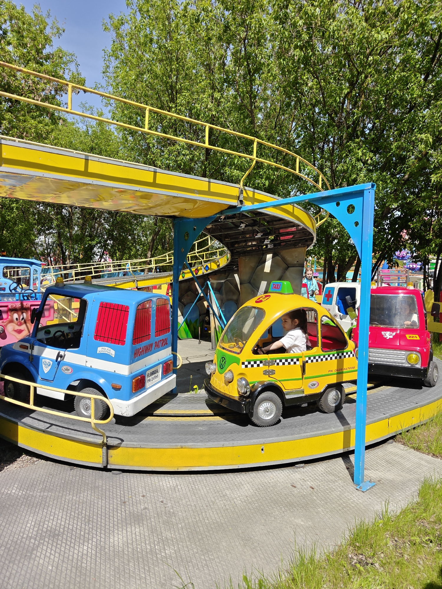 In the city of Khimki, Russia, a carousel features a yellow rotating platform with colorful toy vehicles—a blue "Highway Patrol" car, a yellow "Taxi" with a child inside, and a red car—under a yellow and blue overhead structure. The background shows trees and a blue sky.