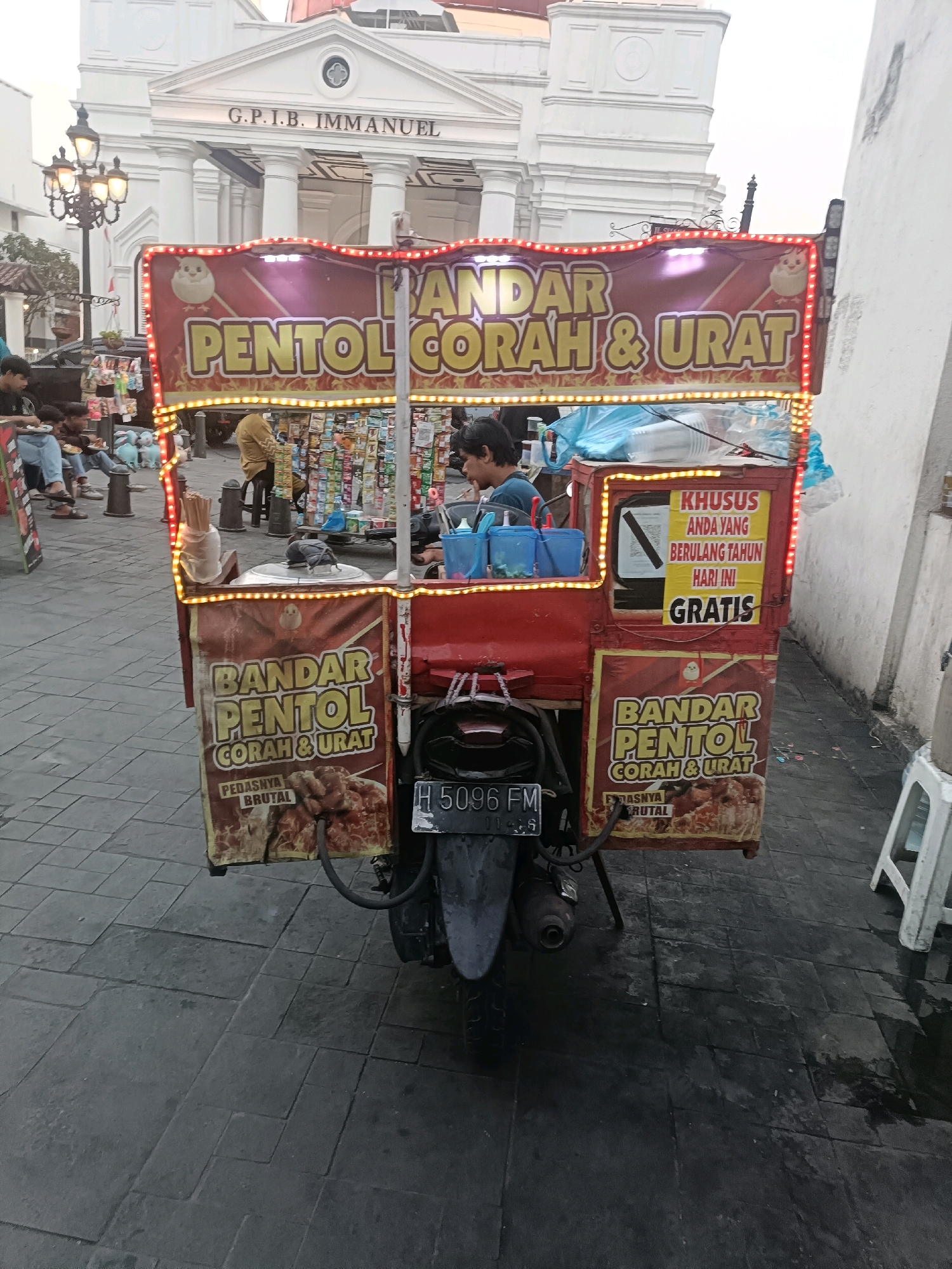 The image shows a food cart, identified as "BANDAR PENTOL CORAH & URAT", in front of a white building with the inscription "G.P.I.B. IMMANUEL". The food cart is decorated with red and yellow lights and features various signs in Indonesian. One sign states "KHUSUS ANDA YANG BERULANG TAHUN HARI INI GRATIS" which translates to "SPECIAL FOR YOU WHOSE BIRTHDAY IS TODAY FREE". The cart is parked on a paved street, with several people visible in the background, some sitting and eating, and a vendor attending to the cart. The scene appears to be outdoors, possibly in a public space or plaza. The overall impression is a street food vendor operating in a urban setting with a notable architectural background.
