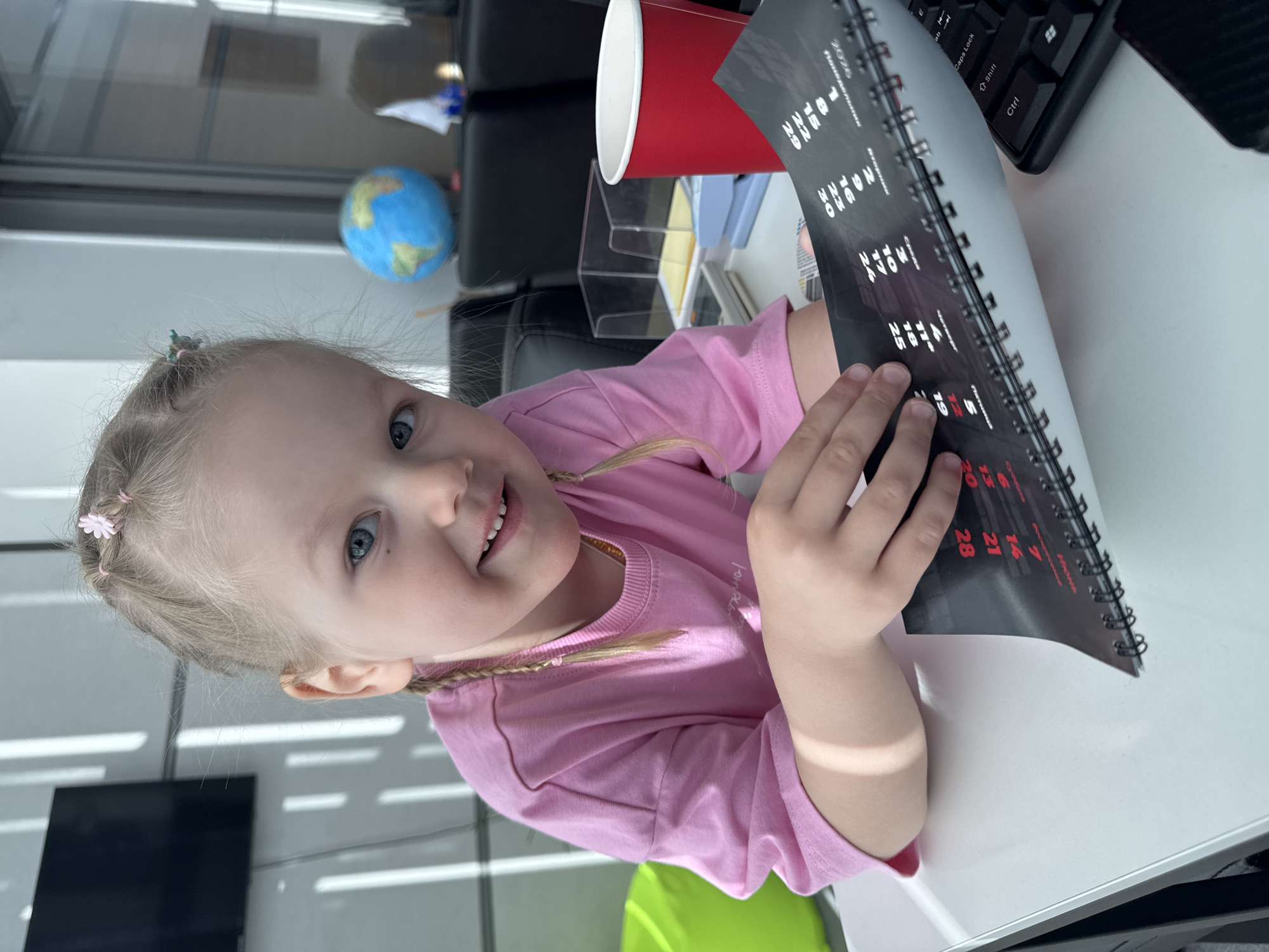 A young, fair-skinned child with blonde hair and blue eyes, likely a girl, is shown smiling and looking directly at the camera. She has small braids on the sides of her head adorned with pink and teal flower-shaped hair clips. She is wearing a pink long-sleeved t-shirt and is seated at a white desk in an indoor office setting in Perm, Russia.
The child is holding a black spiral-bound calendar or planner, which features white and red numbers against a dark background. Visible text on the calendar includes "2026", "Понедельник" (Monday), and "Кино!". Dates such as "1, 8, 15, 22, 29", "2, 9, 16, 23, 30", and other single and double-digit numbers up to 28 are clearly legible.
On the desk, to the child's right, is a red paper cup. Partially visible behind it is a clear acrylic desk organizer. A small blue and green globe is visible further in the background on the desk. A black computer keyboard, with "Caps Lock", "Shift", and "Ctrl" keys visible, is also on the desk. The background features what appears to be a large window or glass partition with horizontal lines, suggesting an office environment. A black office chair is partially visible, and a vibrant lime green chair is seen beneath the desk. The child appears happy and engaged, suggesting a moment of play or curiosity in a workspace.