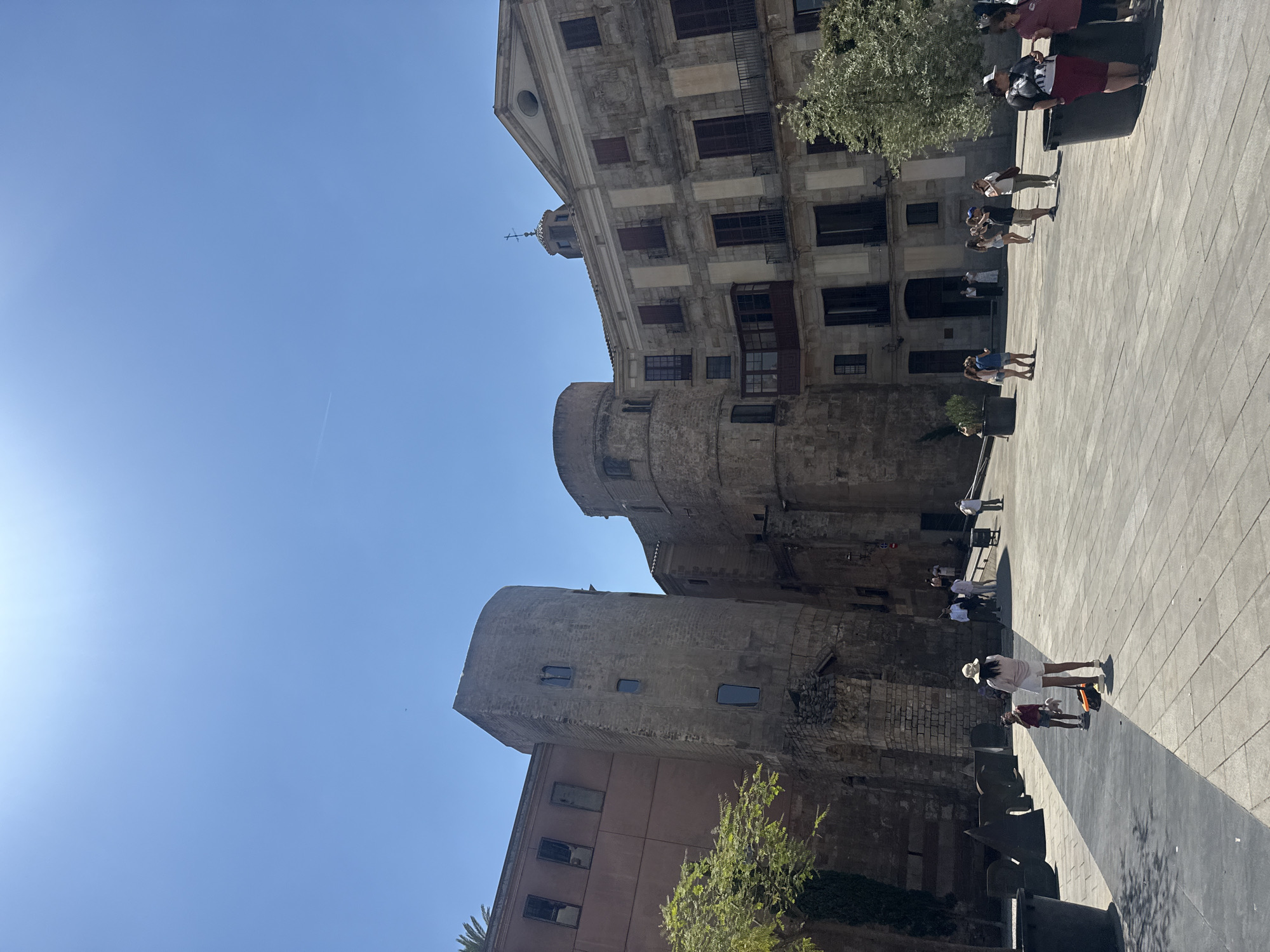 This media file shows a view of a historical building complex in Barcelona, Spain, on a sunny day. The scene is dominated by ancient stone structures, including a large cylindrical tower and a multi-story building with a red facade. Above these, a grander, older stone building with a triangular pediment and a small tower or spire is visible against a clear blue sky. Several people are walking and standing in a tiled plaza in front of the buildings, enjoying the bright sunlight.
The time of day appears to be midday or early afternoon, as indicated by the strong shadows cast across the plaza. The weather is clearly fair and sunny, with a cloudless sky. The architectural style suggests a historical European location, and the context provided specifies Barcelona. The people are casually dressed, suggesting they are tourists or locals enjoying the public space. One person in the foreground, a woman wearing a white hat and a light-colored dress, is facing away from the camera. Another individual, a child, is lying down near her. Other people are seen in groups or individually further back in the plaza. The overall mood seems relaxed and pleasant, with people engaging in leisurely activities in a historically significant environment. No visible text is present in the image.