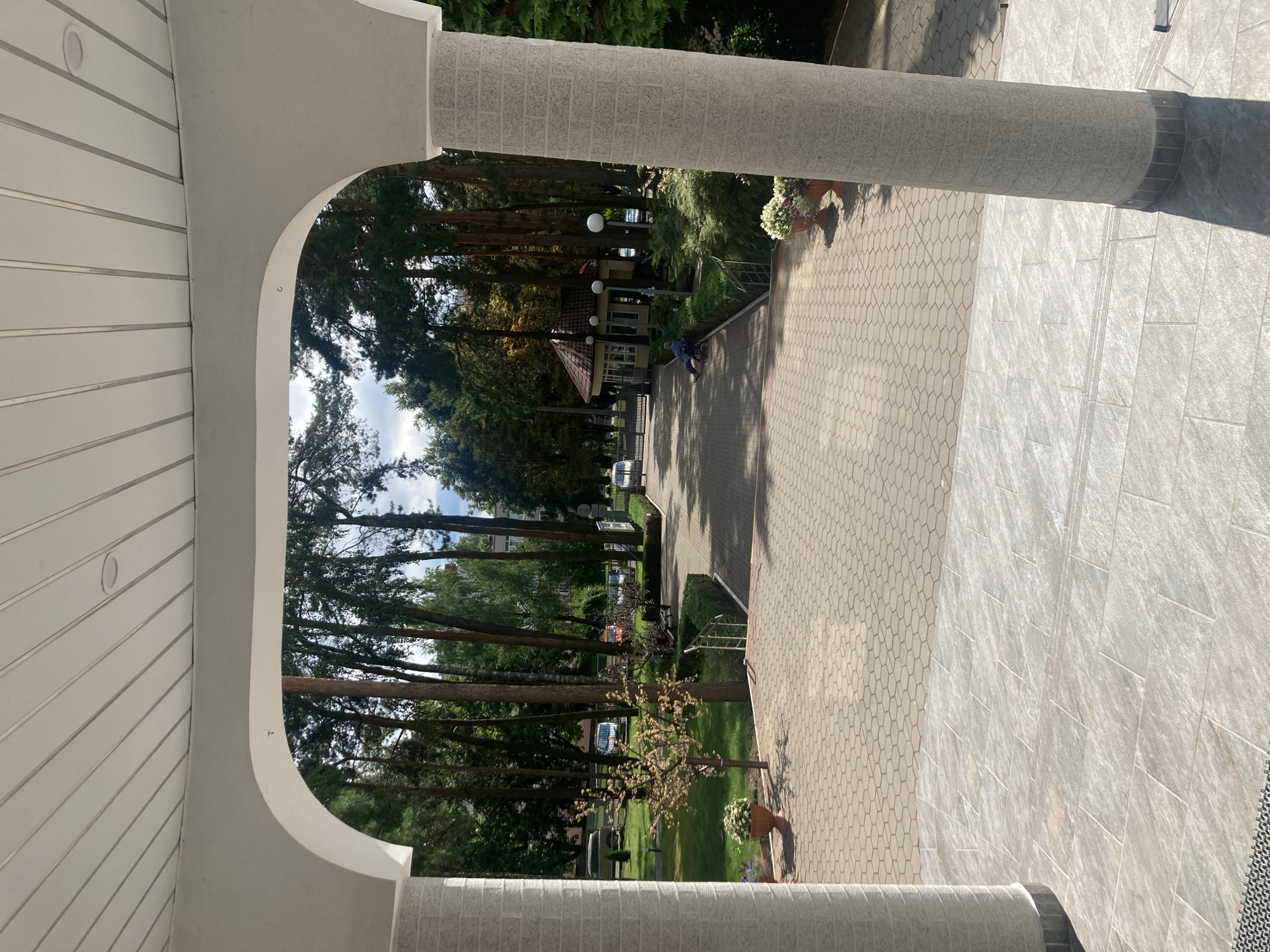 The image captures an exterior view from under a white arched structure, looking out onto a tree-lined pathway and manicured grounds. The foreground features a tiled patio leading to a hexagonal paver pathway. To the left, a textured white column stands, and a large potted plant with white flowers is visible. The pathway extends into a park-like setting with numerous tall trees, green grass, and neatly trimmed hedges.
In the middle ground, a small, octagonal building with a tiled roof and windows is situated. Two round lampposts stand in front of it. A person in a blue shirt is seated on a bench to the left of the building. Several parked cars are visible further down the pathway. A metal fence and gate are also present on the left side of the pathway.
The background shows more trees and the facade of a building in the distance. The sky is partly cloudy, and the overall impression is of a serene, well-maintained outdoor area. This scene is consistent with the grounds of the Baranavichy State University in Baranavichy, Belarus, known for its park-like campus and architectural elements.