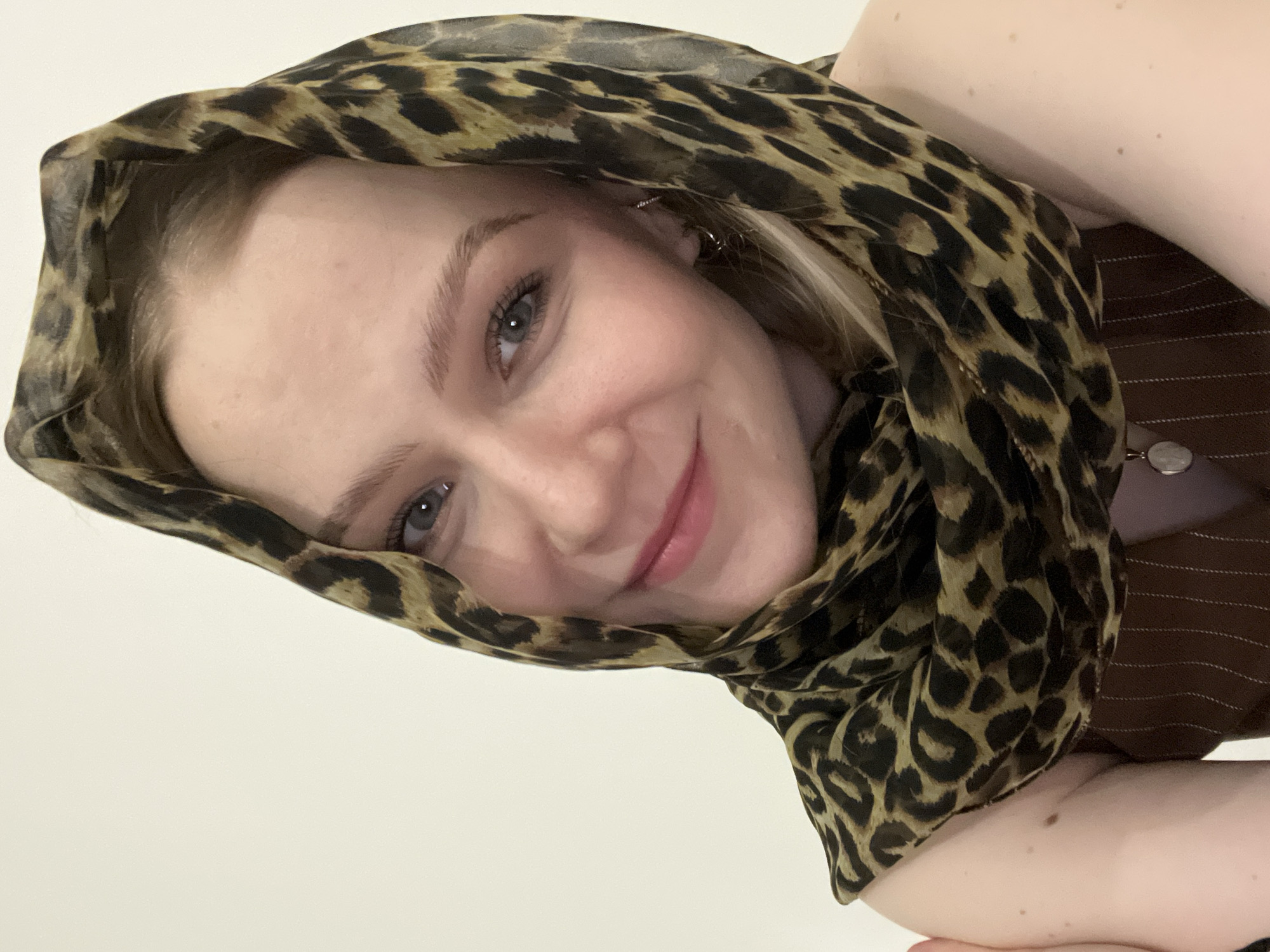 A young woman is pictured indoors. She is wearing a leopard print scarf wrapped around her head. She is also wearing a brown pinstripe vest. Her arms are visible in the bottom right corner. The background is white. There is no evidence in the image to suggest a location in Khimki, Russia.
