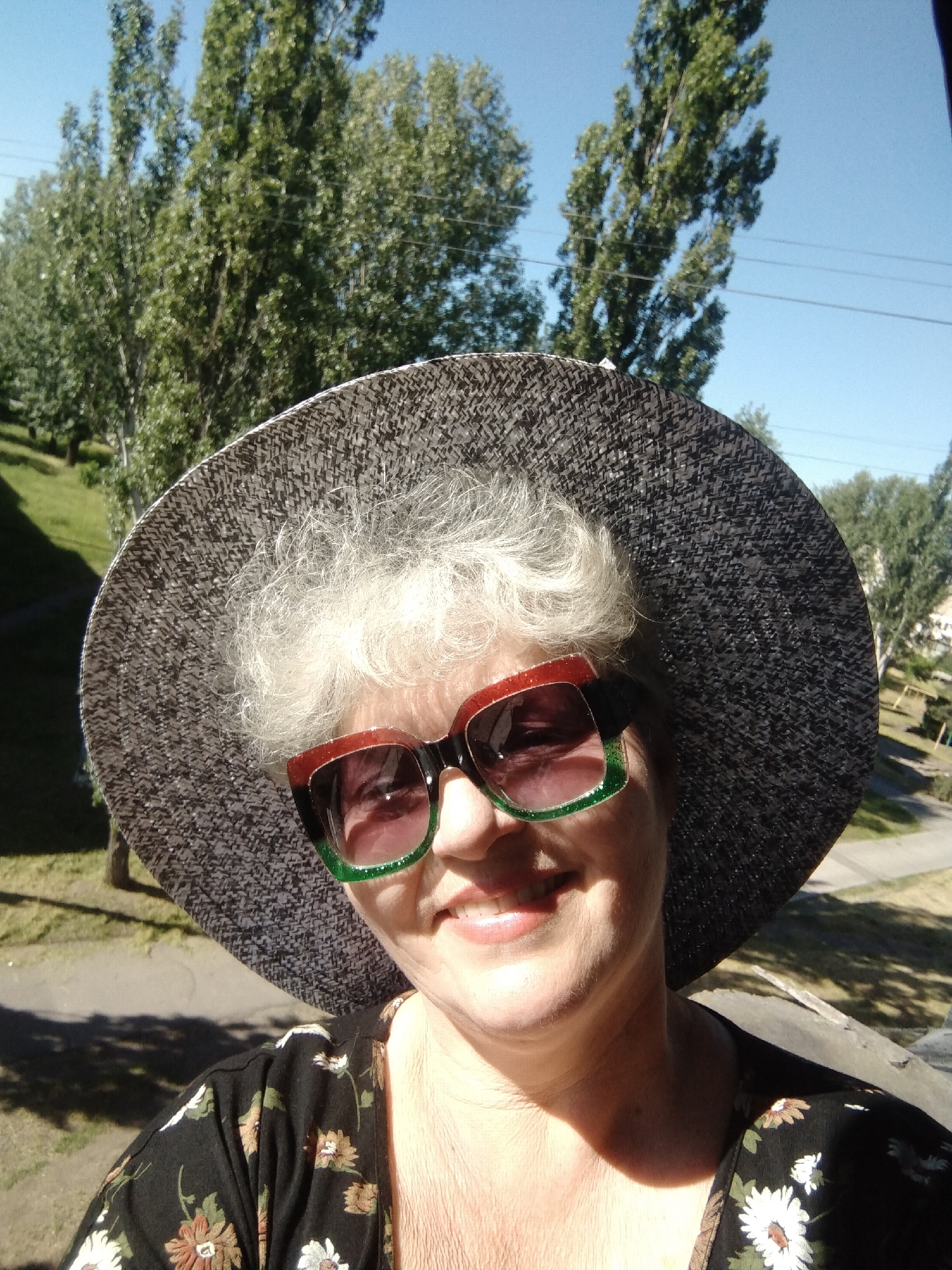 A close-up shot features an elderly woman in a large, dark straw hat with a textured surface. She is smiling, displaying her teeth, and wearing sunglasses with red and green frames. Her hair is short, curly, and white. She is wearing a dark floral shirt with a pattern of white and brown flowers.
The background includes green trees under a blue sky. A paved area is also visible. Based on the surroundings, the location appears to be an outdoor setting in Horishni Plavni, Ukraine. The power lines can be seen in the blue sky, which is another detail that is essential.