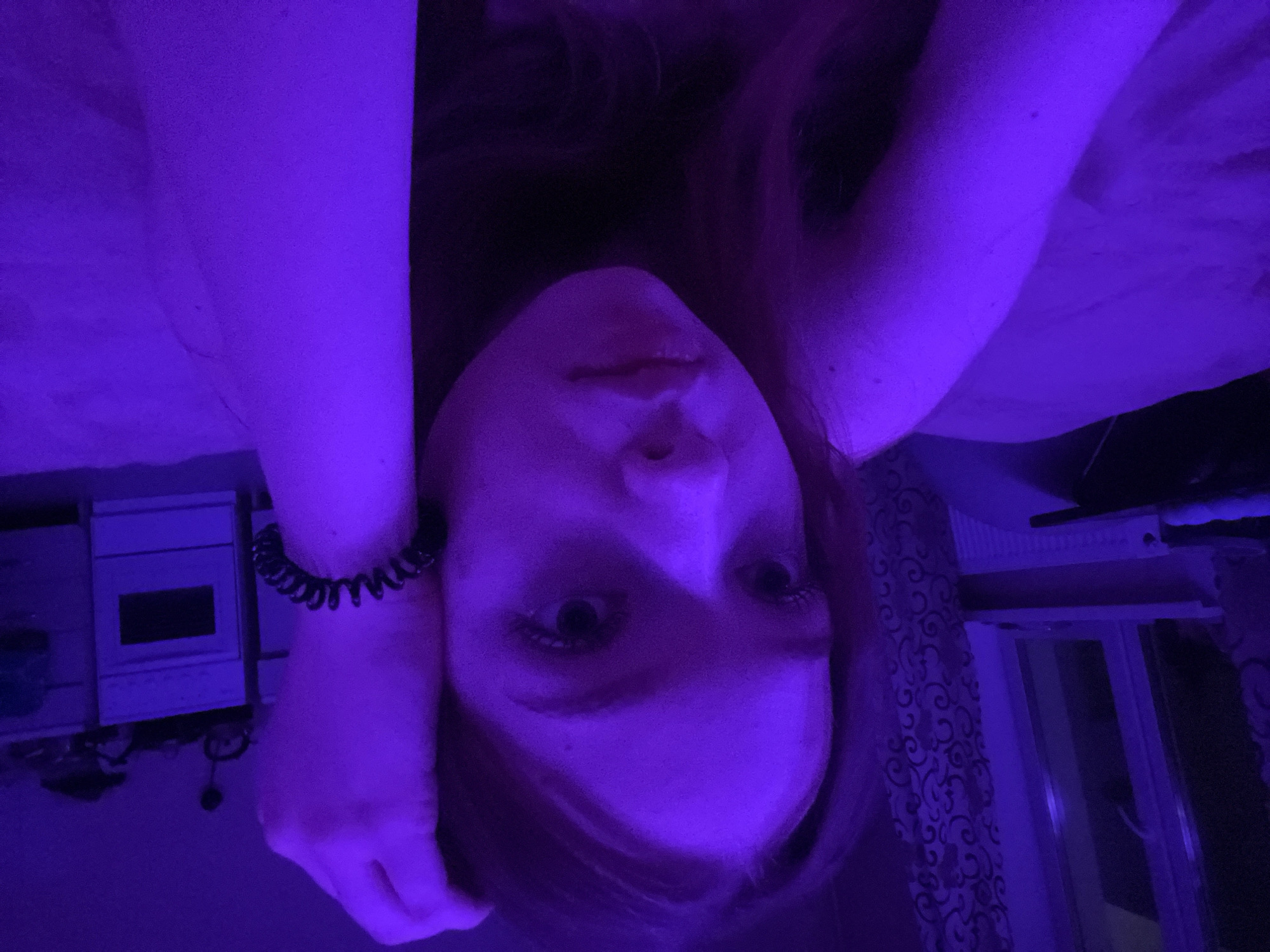 A person lies in bed in a room illuminated by purple light. The person has reddish-brown hair and is looking directly at the camera with their arms raised on either side of their head. The person is wearing a black hair tie on their left wrist. The room includes a bedside table with objects on it, a window with patterned curtains, and other items that are difficult to discern due to the lighting and angle. The location is likely Chaikovsky, Russia.