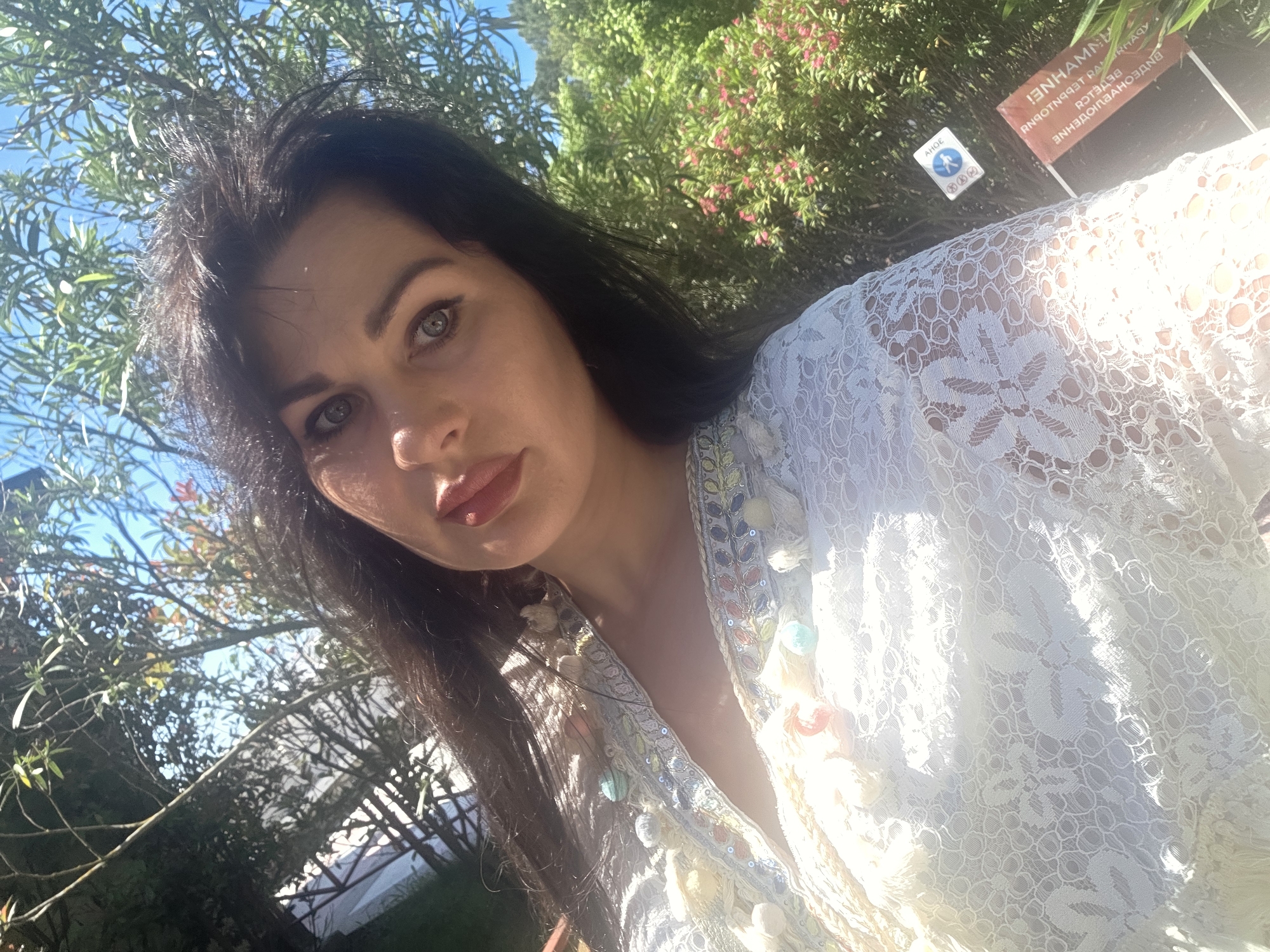 A woman with dark hair and full lips takes a selfie outdoors. She is wearing a white lace garment adorned with colorful beads and tassels. Sunlight illuminates her face and the details of her clothing. Behind her, lush green trees and a bright blue sky are visible. In the background, there are two signs, one small and white with a blue pictogram of a person walking and the other a larger red sign with text. The overall impression is that of a bright, sunny day in a natural setting, possibly a park or garden within the city of Sochi, Russia.