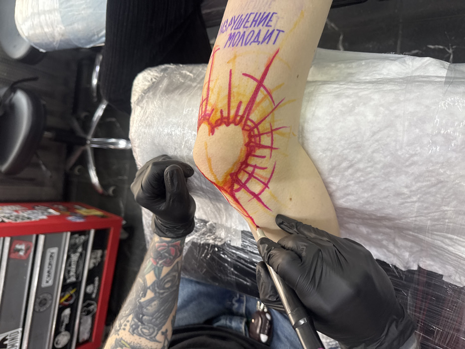 A person's arm, bent at the elbow, is being tattooed. The elbow is encircled by a sunburst design in red and orange ink, with rays emanating outward. Text in Russian, "ИЗВРАЩЕНИЕ МОЛОДИТ," is written above the tattoo design. The tattoo artist is wearing black gloves and working on the lower part of the sunburst with a tattoo machine. A red toolbox with stickers is visible in the background. The arm is resting on a white, disposable barrier.