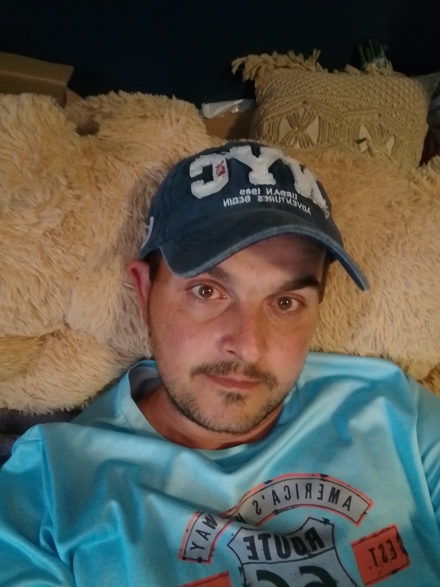 A man wearing a blue baseball cap and a light blue t-shirt is shown from the chest up, lying down. He has brown eyes and a beard and mustache. The background consists of plush, tan-colored pillows and a decorative macrame pillow. The t-shirt has graphics on it. The scene appears to be indoors. There is no information in the image to suggest it is in Wedel, Germany.